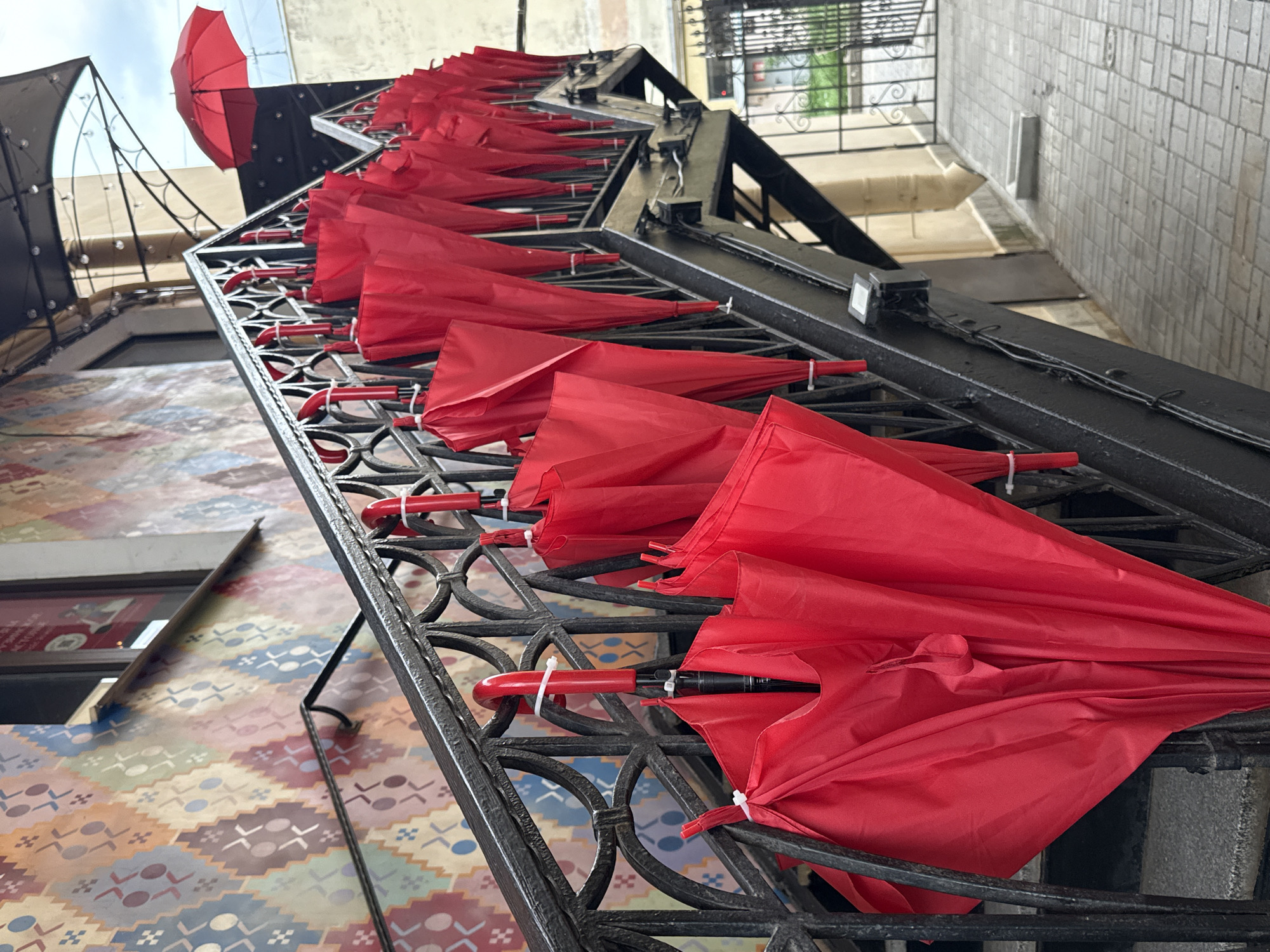 A group of red umbrellas are hanging on a black metal railing along a staircase, in front of a building with a colorful, patterned mural. The umbrellas are closed and tied with white plastic ties. One red umbrella is open and extended above the railing further up the stairs. The scene appears to be outdoors, on a cloudy day. The architecture and the style of the mural suggest a location in Saint Petersburg, Russia.