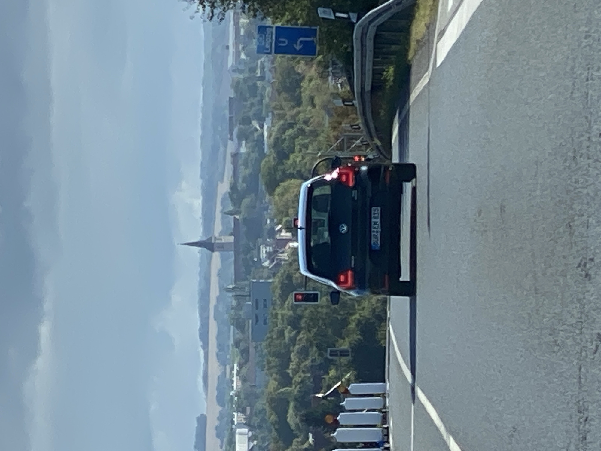 The image shows a street scene in Werther, Germany, on a cloudy day. A dark-colored Volkswagen Polo is driving away from the camera down a paved road. The car has a license plate that reads "BU" followed by some digits and letters, partially obscured. The car's brake lights are illuminated. To the left of the road, there are several white bollards. Further to the left, a traffic light is red. In the background, a town with buildings and trees is visible, with a church spire prominently featured. A blue directional sign with the word "Leipzig" and a U-turn arrow is visible on the right side of the road, indicating a route towards Leipzig. The road appears to be a highway or a main road leading into or out of the town.