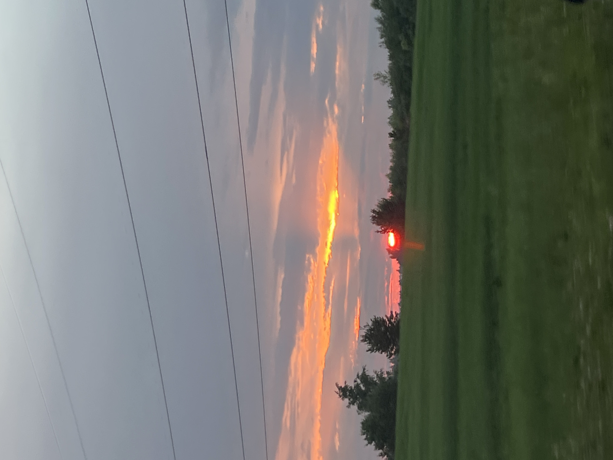 The image depicts a sunset over a rural landscape, likely in the vicinity of Поварёнки, Russia. A vast expanse of green field stretches across the foreground, with a line of trees forming a distant horizon. Above the trees, the sky is a canvas of dramatic colors. A band of vibrant orange and yellow clouds is illuminated by the setting sun, which appears as a bright red orb peeking through a break in the clouds. Faintly visible in the upper portion of the image are power lines, suggesting a proximity to infrastructure. The overall impression is one of natural beauty and tranquility, with the dramatic sunset providing a focal point against the serene rural backdrop. There are no people or animals visible in the image.