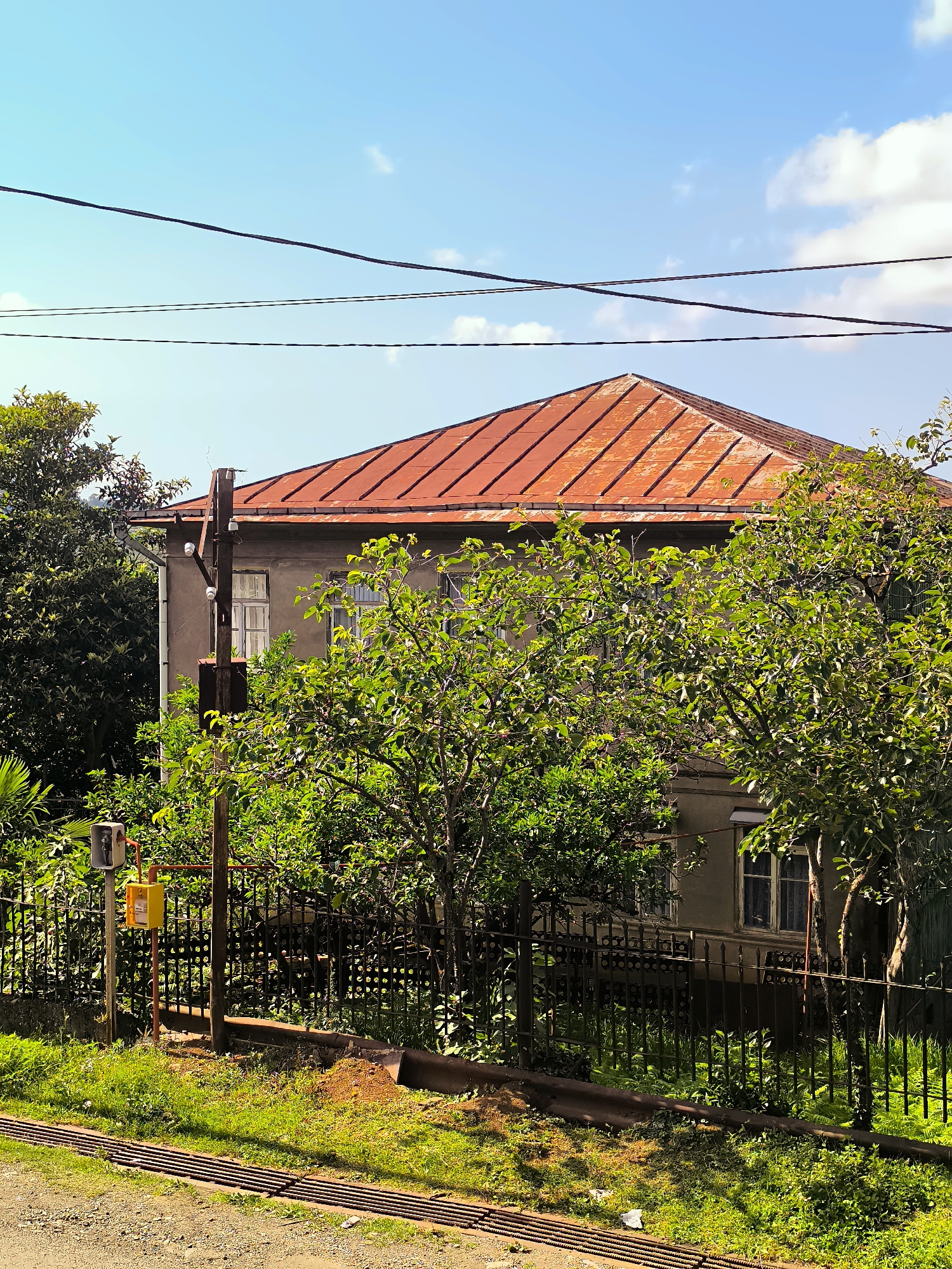An exterior, eye-level, medium shot captures a weathered, two-story house in Erge, Georgia. The house features a rusted, orange corrugated metal roof and light gray walls. Several windows with white frames are visible on the facade, some covered with curtains. In the foreground, a black metal fence encloses a garden with lush green trees and bushes. A wooden utility pole with ceramic insulators stands near the fence, alongside a yellow electrical box and a grey utility meter. Overhead, black electrical wires stretch across the clear blue sky, with a few white clouds scattered in the distance. The ground in front of the fence is covered with green grass and gravel. The scene appears to be a residential area on a sunny day.