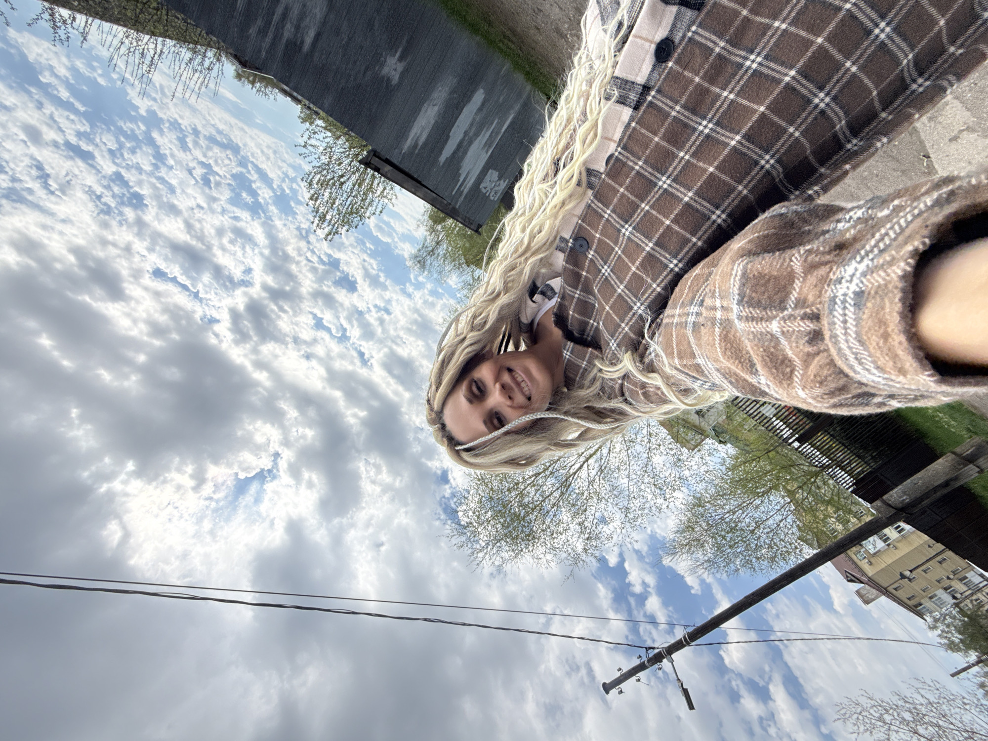 A woman with long blonde braids is taking a selfie outdoors. She is wearing a brown and beige plaid shirt. The background consists of a cloudy sky, power lines, trees, and buildings. Based on the architectural style of the buildings, the location is likely Rusanovo, Russia.