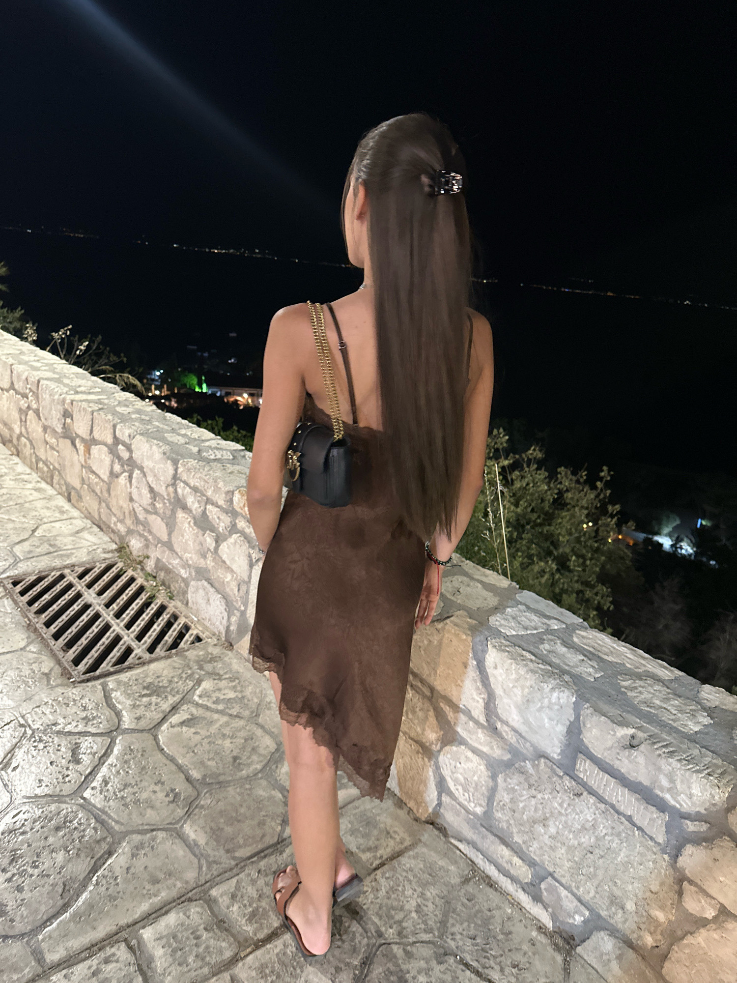 A woman stands facing away from the camera, looking out over a body of water at night. She is wearing a short, brown, slip dress with a lace trim and a small black purse with a gold chain strap. Her long, brown hair is pulled back with a claw clip. She is wearing flat, brown sandals. She is standing on a stone pathway next to a stone wall. A metal grate is visible on the pathway. The background is dark, with lights visible across the water, suggesting a town or city. The setting appears to be a coastal area in Kassandra, Greece, given the context provided.