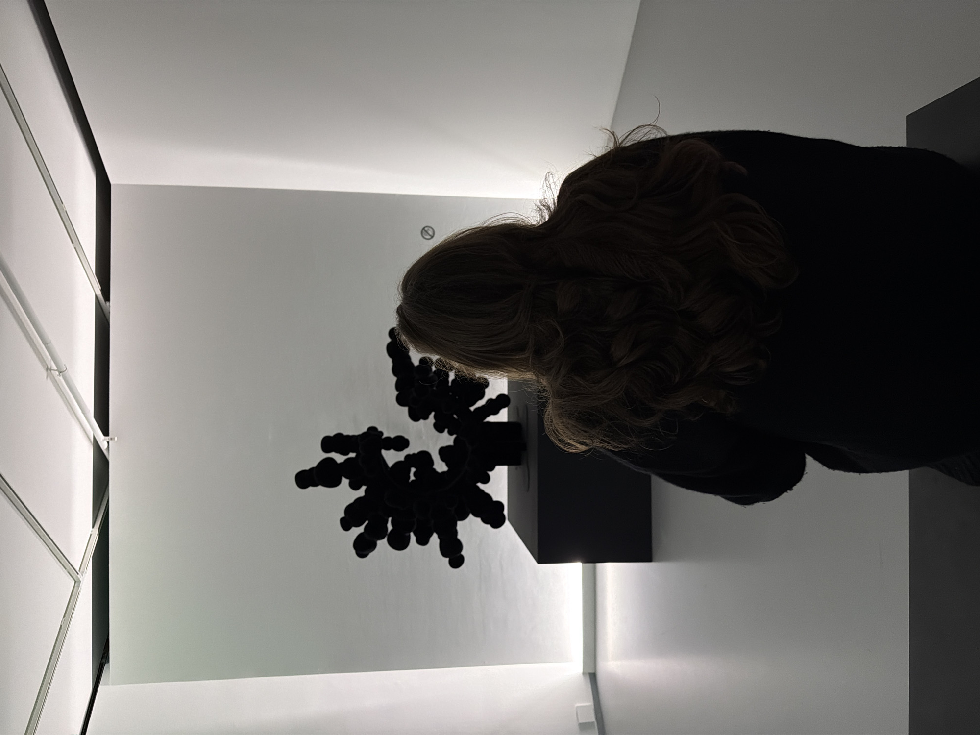 A slightly rotated, low-angle, indoor shot captures a woman with wavy brown hair wearing a dark-colored top, positioned to the right of a dark, abstract sculpture. The sculpture, resembling a cluster of spheres, is mounted on a black cubic base. The scene is set in a white room with geometric paneling visible on the left side, suggesting an art gallery or exhibition space. A circular object, potentially a sensor or indicator, is discernible on the wall near the woman's head. Given the visual style and prevalent art scene, the location might be an art space in a major city, potentially Moscow, Russia.