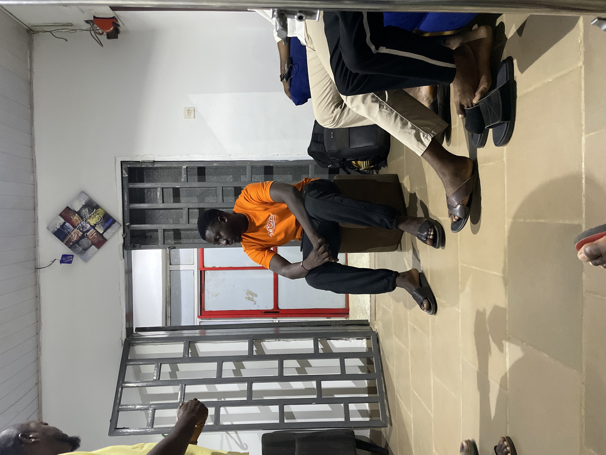 This image, taken indoors in Barakin Akawo, Nigeria, depicts a casual gathering of at least four individuals, likely in the evening or at night due to artificial lighting and prominent shadows. The setting features light-colored walls and a light brown tiled floor.
In the center-left, a man wearing an orange t-shirt with an unidentifiable white graphic and black pants is seated on a low structure, possibly a bench, looking downwards. He wears black slide sandals. Behind him, a metal security gate or window bars are visible. To his right, two other individuals are partially seen, primarily their legs and feet. One wears khaki pants and black and white striped pants, along with black slide sandals. Another has black pants and brown leather sandals. A black backpack rests on the floor near their feet. A fourth person, wearing a yellow shirt, is partially visible in the bottom left foreground, also looking down.
Notable objects include a colorful framed artwork on the wall to the left of the barred opening, an exposed electrical fixture on the ceiling in the top-left corner, and a small white electrical outlet on the wall. The overall mood appears relaxed and informal. No clearly legible text is visible.