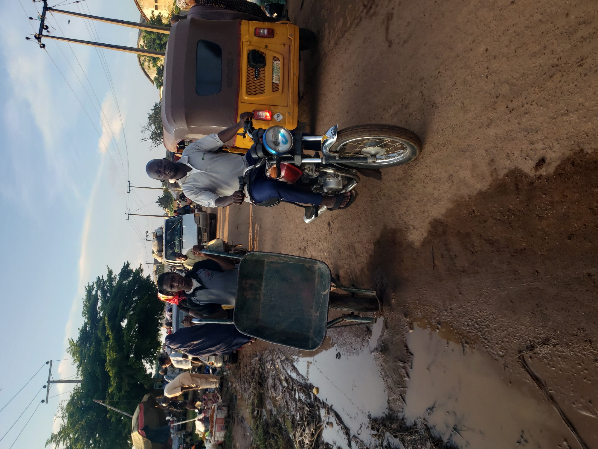 In Kakuri Gwari, Nigeria, two individuals are captured in a street scene. On the left, a young boy, wearing a grey and black shirt, pushes a green wheelbarrow with metal handles. The wheelbarrow is empty and positioned on a muddy, uneven dirt road. Behind him, several people are gathered around market stalls, with one person in a blue garment standing near the wheelbarrow. To the right, a man wearing a light grey polo shirt and dark blue trousers rides a red and chrome motorcycle. He is looking forward with his eyes closed. The motorcycle's front tire is visible, and a yellow auto-rickshaw with a brown canopy is situated behind him. The environment appears to be a marketplace with visible power lines and trees in the background, under a bright sky with scattered clouds.