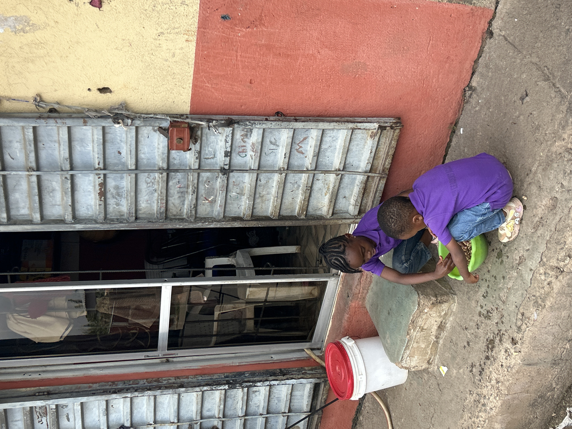 Two children in purple shirts and blue jeans are playing with a green bowl of beans on a concrete surface near a building. The building has a red and yellow wall, metal sliding doors, and a window showing interior contents. A white bucket with a red lid sits nearby. The setting is likely in a residential or commercial area of Jos, Nigeria.