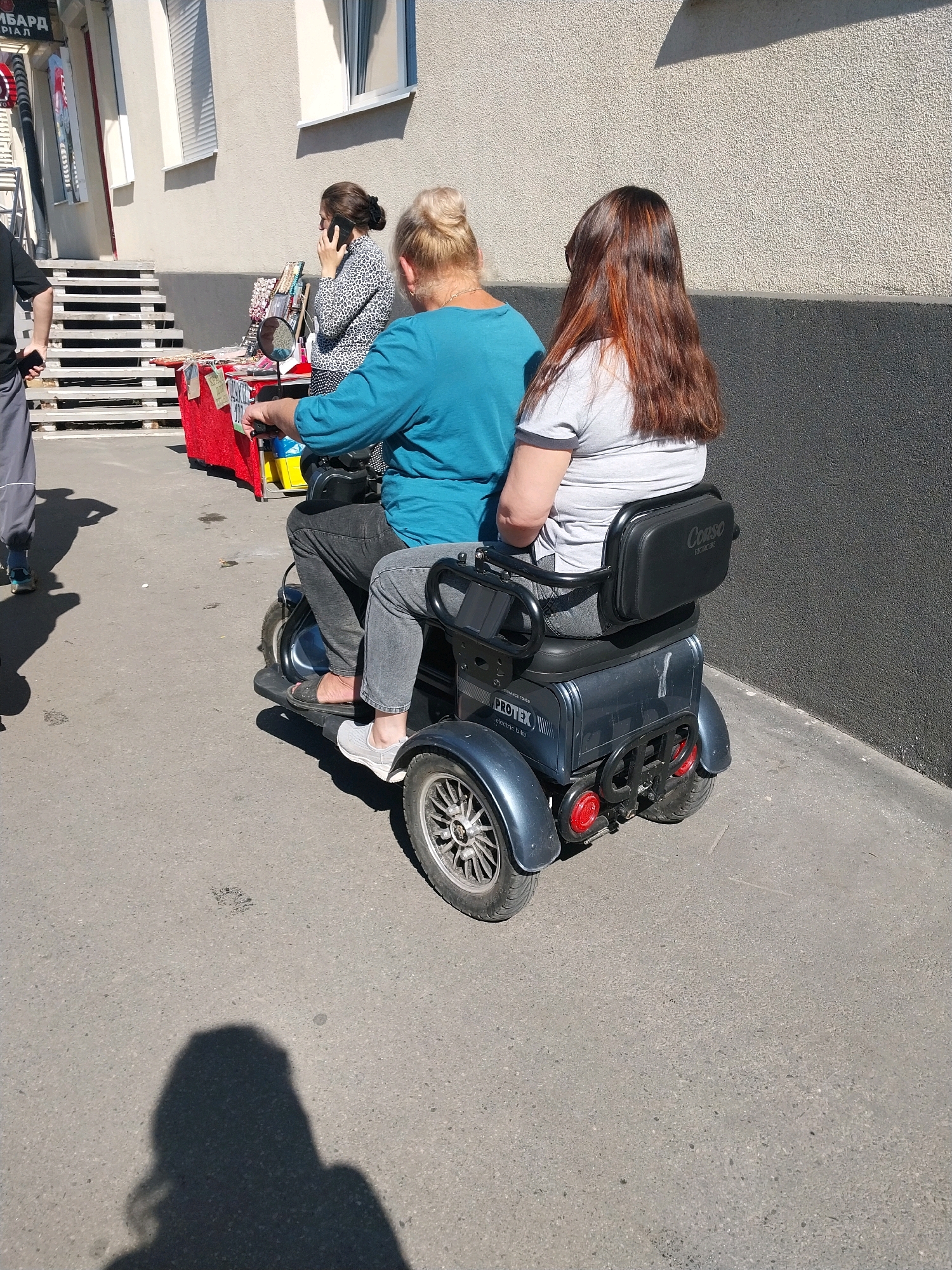 Two women are riding on a three-wheeled electric scooter. The woman in the front is wearing a turquoise long-sleeved shirt and gray pants. The woman in the back is wearing a gray t-shirt with a black trim and has long brown hair. The scooter has a black seat with a headrest that has the logo "Corso Electric Bike" printed on it, and a label "PROTEX electric bike" on the side. They are on an asphalt surface in front of a building with a textured, light-colored facade and dark gray lower wall. To the left of the scooter, a woman in a leopard-print shirt is standing and talking on a mobile phone next to a red stall displaying various items, possibly for sale. Behind the stall, there are wooden stairs. The image appears to have been taken in daylight, casting shadows on the ground. The scene is in Dnipro, Ukraine.