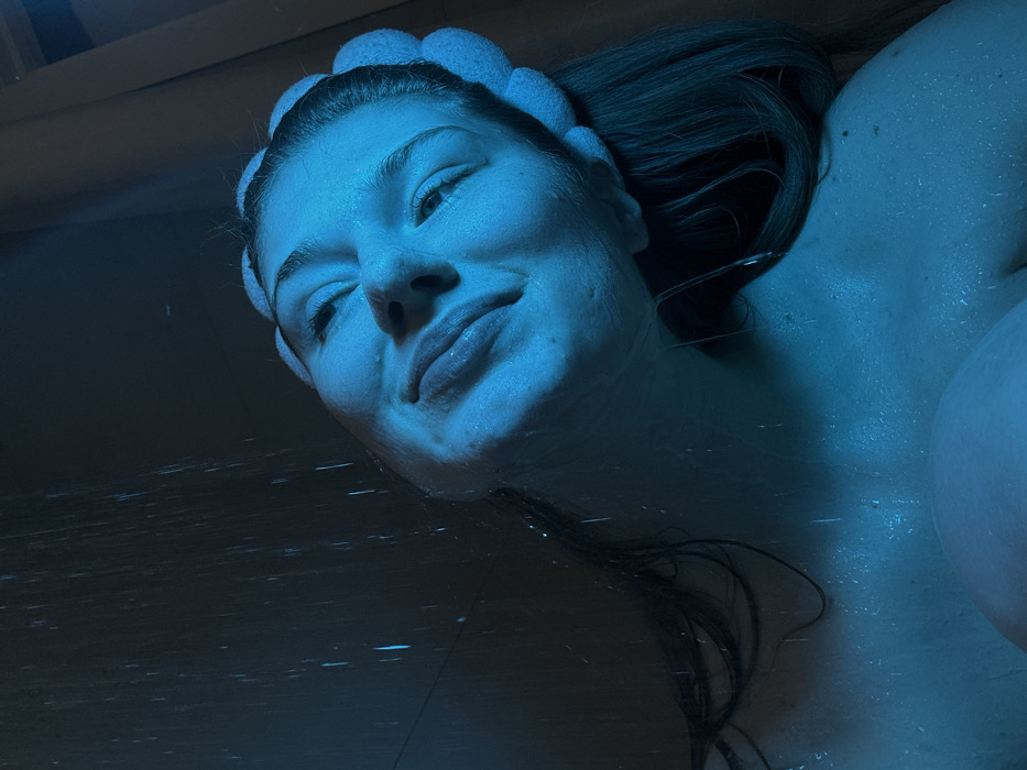 A close-up image depicts a young adult female showering under a blue light. Her head is tilted upwards, eyes partially squinted, and lips slightly parted as water streams down onto her face and upper body from an unseen source above. She wears a light-colored, possibly white or cream, fluffy head-band pushing her dark hair back from her forehead. Water droplets are visible on her wet skin and hair. The immediate background is dark and indistinct, consistent with an enclosed shower environment. No specific identifying features of the city of Kazan, Russia, are discernible within the image itself; however, it is presented as taking place within that city.