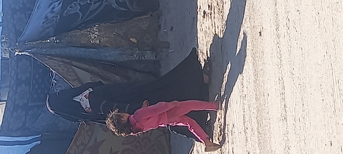 The image depicts two individuals, an adult and a child, walking on a light-colored, dusty ground in Khan Yunis, Palestinian Territory. The adult, positioned behind and slightly to the right of the child, wears a full black abaya or niqab, with a visible white garment featuring a red floral pattern underneath. The adult's right foot, clad in a sandal, is visible. The child, a young individual with dark, curly hair, wears a complete pink tracksuit and appears to be wearing light-colored sandals or walking barefoot. The child is walking slightly ahead of the adult, with the left leg extended forward. Behind the figures, multiple makeshift shelters are visible, constructed from various tarpaulins and fabric materials, some appearing dark with patterns, others lighter. Strong sunlight casts distinct shadows of both figures onto the ground, indicating an outdoor daytime scene.