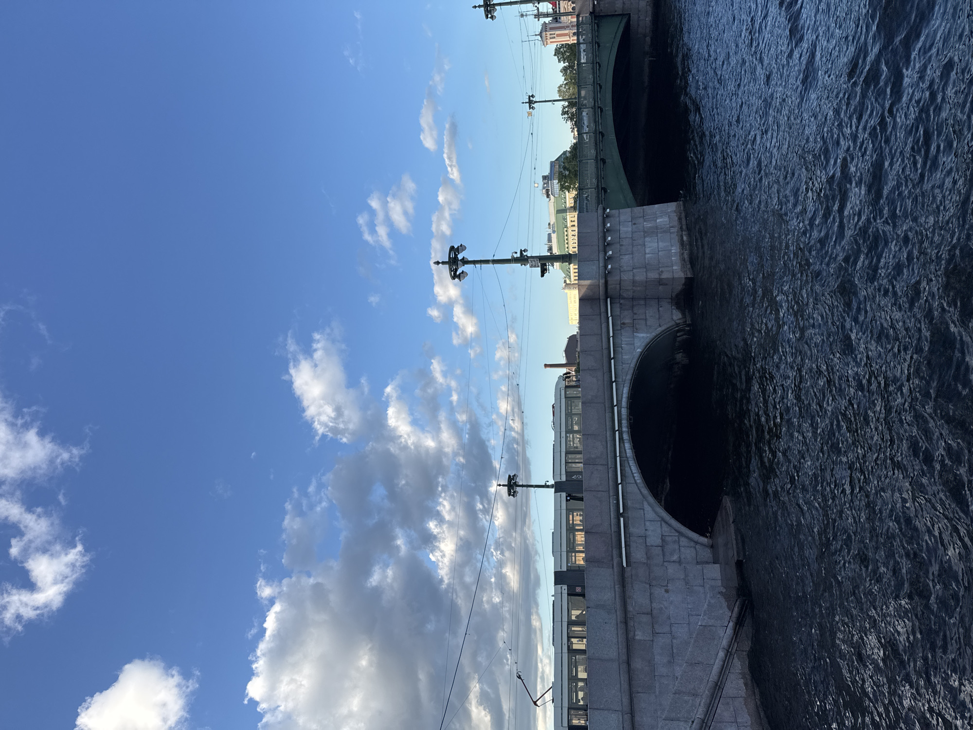 The image depicts a canal in Saint Petersburg, Russia, under a blue sky with scattered clouds. Two bridges span the canal. The foreground shows dark, rippling water. The bridge in the foreground is made of stone and has a large arched opening through which the water flows. Above this bridge, a modern tram with multiple carriages travels on tracks, silhouetted against the sky and clouds. The second bridge, visible to the right and further back, is green and also has an arched opening. Streetlights with multiple lamps are positioned along the bridges. Buildings are visible in the background, including structures with green roofs and ornate architecture. The overall scene captures a typical urban canal view in Saint Petersburg, with a blend of historic bridges and modern transportation.