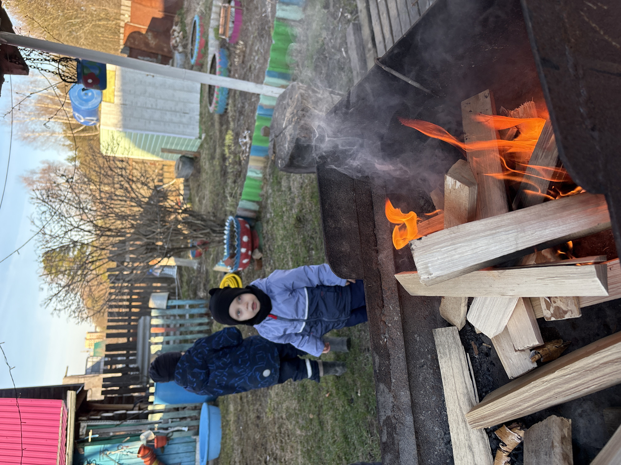 A vibrant outdoor scene depicts two young children standing near a metal brazier with an active fire, set in what appears to be a backyard or garden. The image is captured in bright daylight, suggesting a clear day, possibly in spring or autumn given the children's jackets and the sparse trees.
In the foreground to the right, a black metal brazier holds a lively fire with bright orange flames and rising grey smoke, fueled by several pieces of light-colored wood. To the left of the brazier, two children are present. The child closer to the camera wears a black beanie with ear-like features, a light purple and dark blue winter jacket, and dark blue pants with grey boots, looking towards the viewer with a calm expression. Slightly behind and to the left of them, a second child in a dark blue patterned jacket and dark pants also observes the scene.
The background reveals a rustic yard with patches of dry grass and several bare-branched trees under a clear, bright sky. A wooden fence extends across the middle ground. Various elements suggest a home garden or dacha setting: colorful painted tires are arranged as decorations or planters, and structures like a light-colored building with siding and a small shed with a red corrugated roof are visible. Blue plastic barrels or containers are also scattered in the yard.
The overall atmosphere is one of relaxed outdoor activity, perhaps a family gathering or a simple day in the yard. The children's attire indicates cool weather, suitable for an outdoor fire. There is no visible text in the image. The setting is consistent with a backyard in Chaikovsky, Russia.