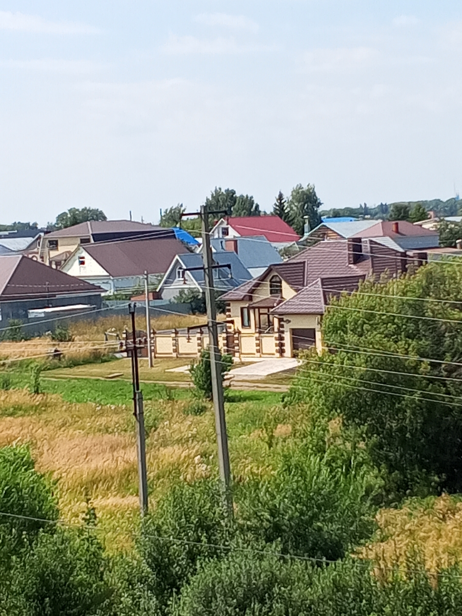 The image shows a residential area in Chistopol, Russia, with several houses visible. The houses have diverse roof colors, including brown, red, and blue. There are utility poles with wires running through the scene, indicating the presence of electricity infrastructure. Vegetation, including green trees and dry grass, is abundant, suggesting a rural or semi-rural setting. The sky is clear with some clouds. The houses appear to be of modern construction, with some featuring garages and decorative elements. The overall impression is that of a quiet, sunny day in a suburban neighborhood.