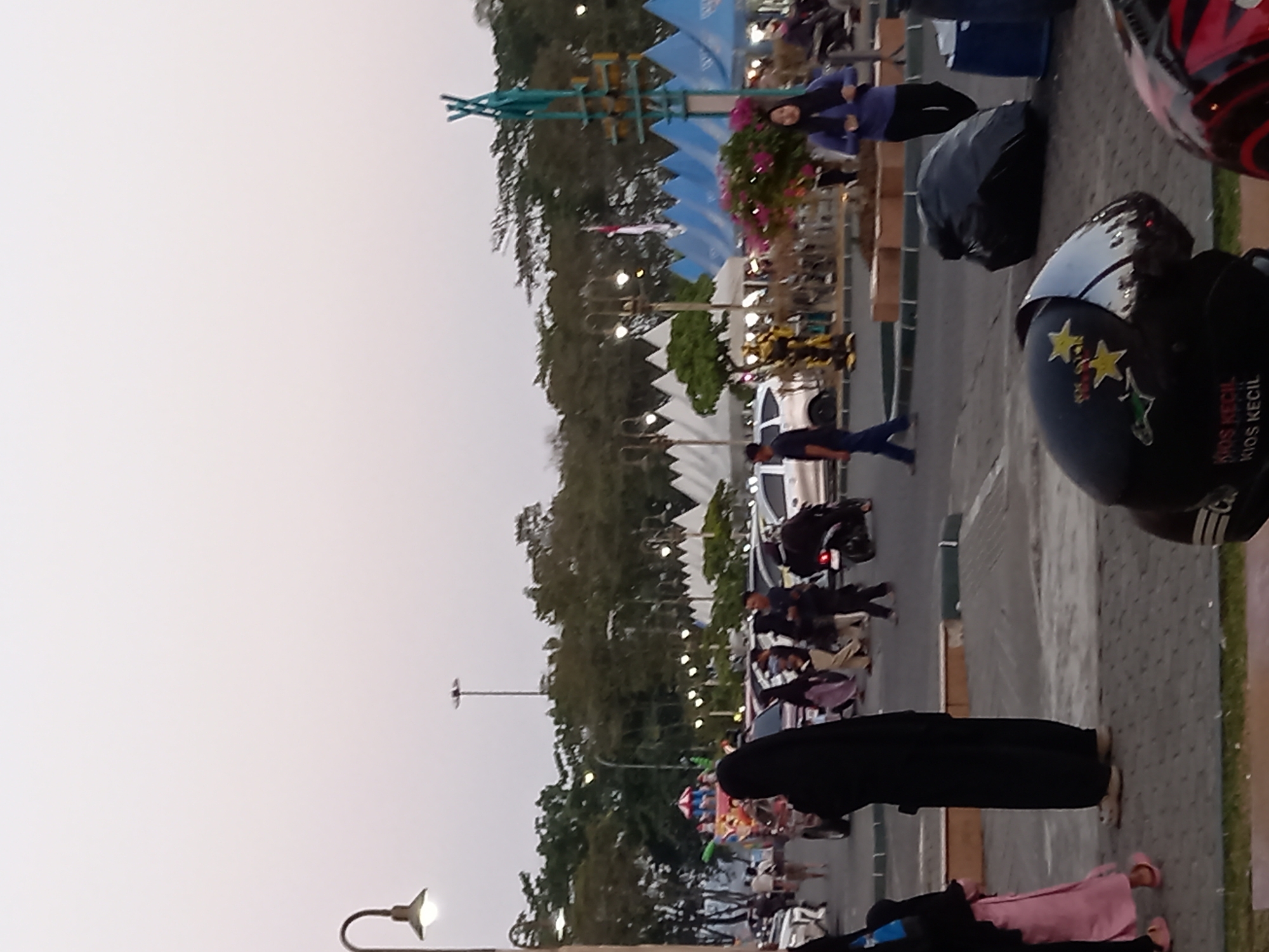 The image captures a street scene in Samarinda, Indonesia, likely in a public gathering area. In the foreground, two motorcycle helmets are visible, one black with star decals and text that reads "KIOS KECIL KIOS KECIL," and another partially visible red and black helmet. In the middle ground, several individuals are walking. A person wearing a black abaya is prominently featured, along with a child in a pink outfit. Other people in modest clothing are also present, some walking in groups. Behind them, various vehicles, including cars and motorcycles, are parked or moving.
The background reveals a lively environment with multiple tents, suggesting a market or event. A tall street lamp is visible on the left. Further back, trees form a dense line, with a flagpole and a large structure that appears to be a tall, thin statue or monument in a teal color. There are also decorative plants with pink flowers. The overall atmosphere appears to be that of an outdoor public space with a festive or commercial activity taking place, possibly in the evening given the lighting conditions.
