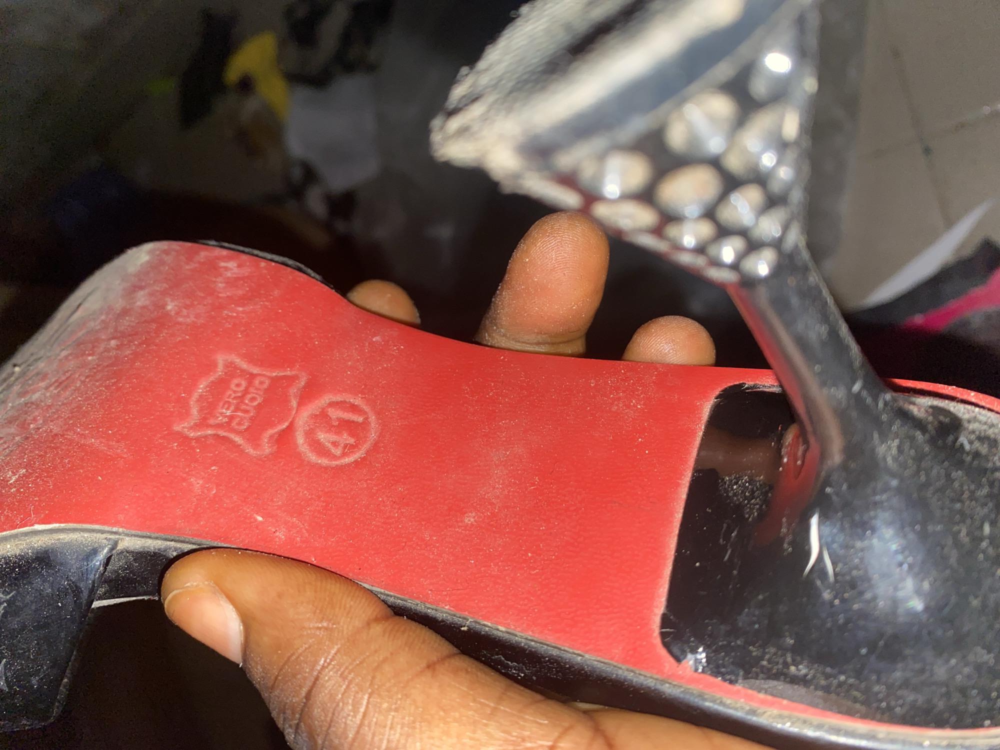 The image shows a close-up of the sole of a red high-heeled shoe, held by a person's thumb and index finger. The sole is red and features a embossed logo with the text "VERO CUOIO" and the number "41" inside a circle. The shoe appears to be dusty. The background is out of focus and dark, with hints of other objects and possibly clutter. The scene does not provide any specific information about the city of Jauro Yinu, Nigeria, or any other urban context.