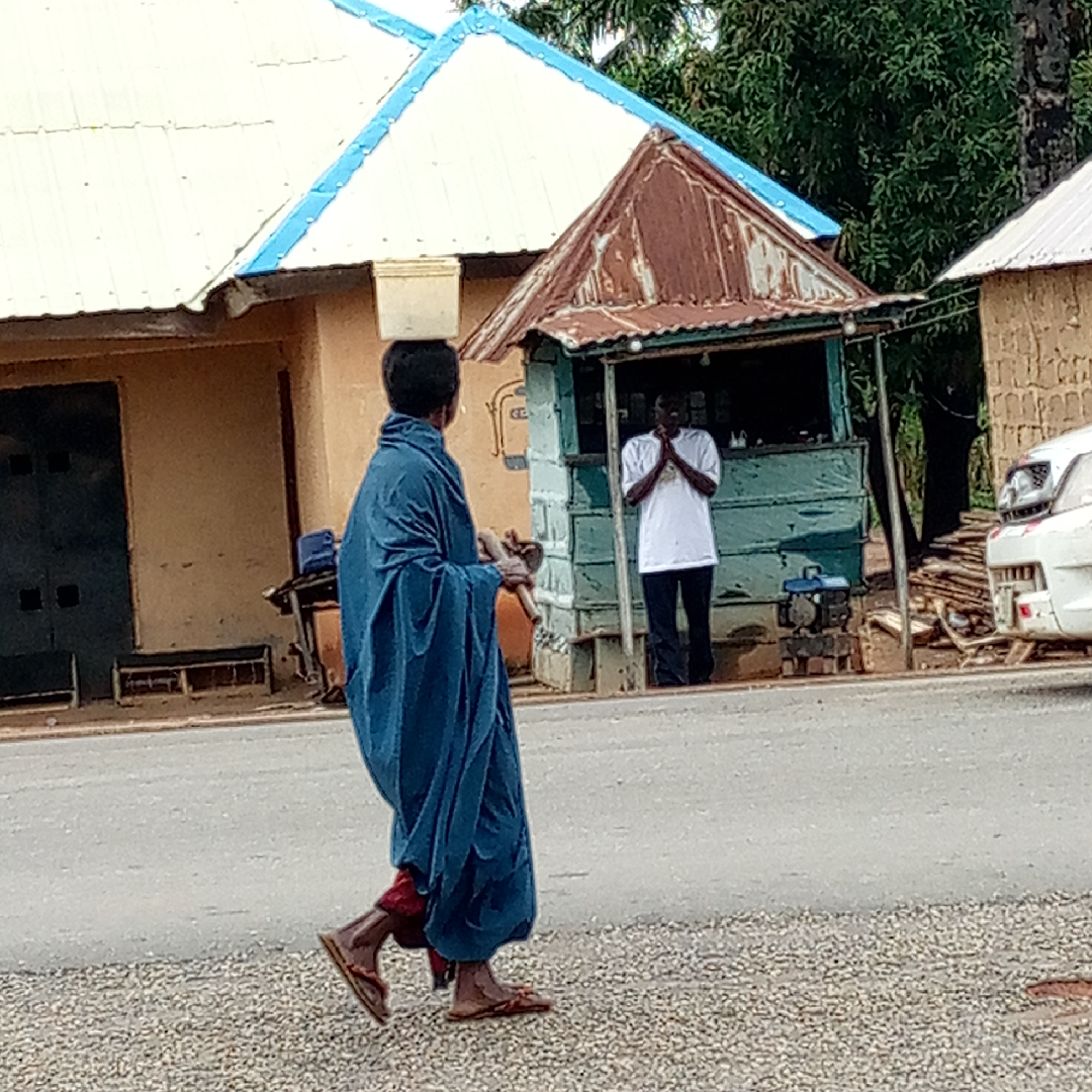 A man in a blue robe and sandals walks along a road in Mararaba, Nigeria, carrying a white bucket on his head and a brown object in his left hand. Behind him, a man in a white t-shirt and dark pants stands in front of a small, dilapidated blue structure with a rusty roof. A generator sits on the ground near the structure. To the right, a white car is partially visible, parked near a pile of wood. Buildings with white and blue roofs are visible in the background. The road surface is asphalt, with a gravel edge on the side where the man in the blue robe is walking.