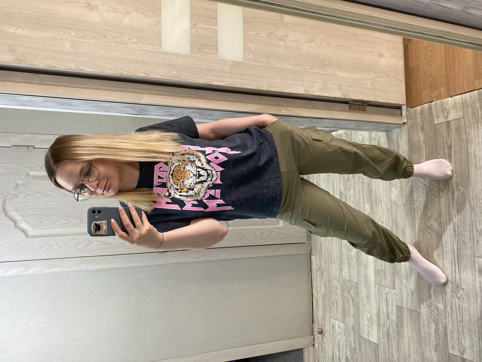 A woman is taking a selfie in a room with wooden laminate flooring. She is wearing glasses, a dark gray t-shirt with a tiger graphic and pink text, and olive green cargo pants. She has on light pink socks. She is holding a gray smartphone with a sticker on the back. A light-colored wooden door is visible behind her and to her right, while a light-colored surface, possibly a wall or another door, is seen to her left. The hinges of the door are visible. The location is Chaikovsky, Russia.