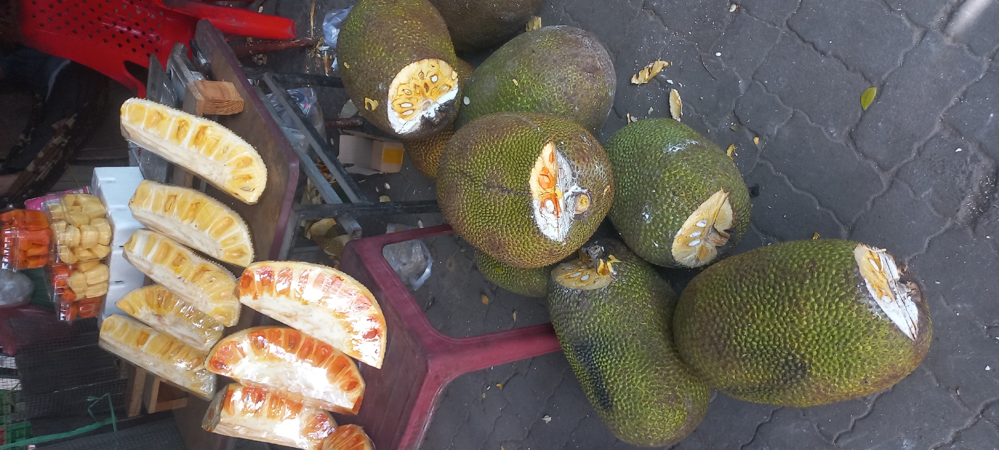 This is a photograph taken at a street-side fruit stand in Da Nang, Vietnam. The foreground features a collection of whole, unripe jackfruits with their characteristic spiky green rinds scattered on a paved surface. Several of these jackfruits have been cut open, revealing their fibrous interior and yellow flesh.
In the midground, on a red plastic table and a metal table with a red chair, are several cut sections of ripe jackfruit. These pieces are arranged and wrapped in plastic. Behind the fruit, there are clear plastic containers filled with more cut jackfruit pieces, some appearing very ripe and golden. The background is blurred but suggests a bustling market environment with other stalls and goods. The overall impression is of fresh produce being prepared and sold outdoors.