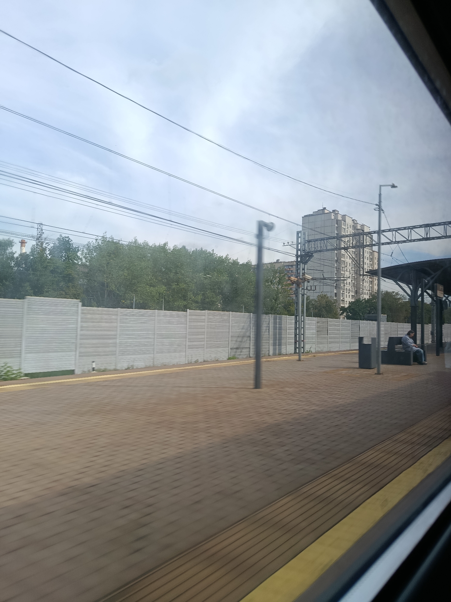 A side view from a moving train shows a pedestrian platform at a railway station in Reutov, Russia. The platform is paved with brown rectangular tiles and has a yellow tactile warning strip along its edge. To the left, a long, gray concrete sound barrier wall separates the platform from a line of green trees and bushes. In the distance, a tall, white residential building with many windows dominates the skyline, along with other buildings and a large chimney with a red top. Overhead, power lines are strung between poles. On the platform, a person is seated on a gray bench under a canopy supported by metal pillars. The sky is overcast with gray clouds.