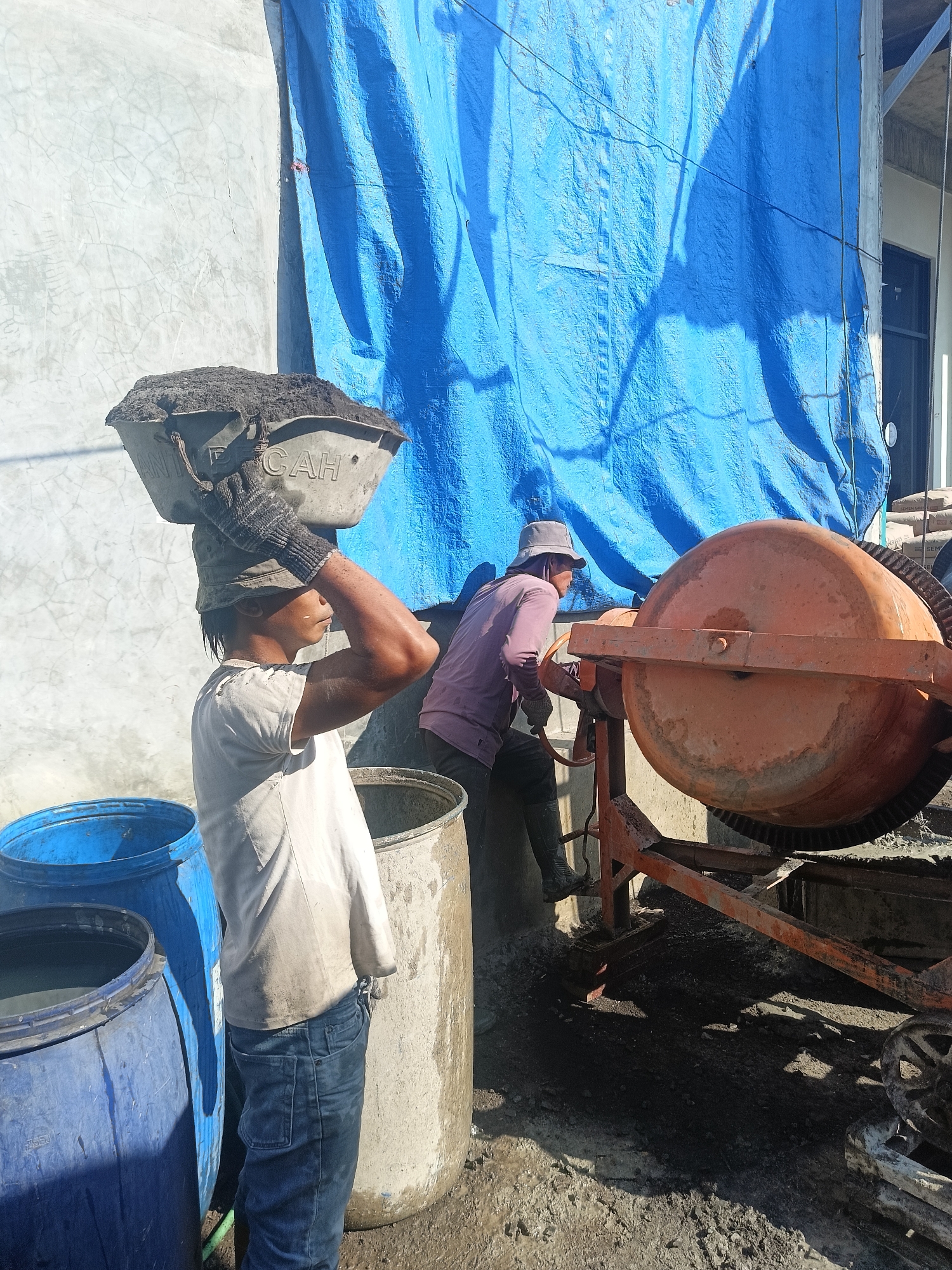 In the city of Gedangan, Indonesia, on a construction site, two men are working with a cement mixer under a blue tarp. The man on the left, wearing a white t-shirt, jeans, and a hat, balances a basin filled with dark material on his head. He is positioned near several large blue and white barrels. The man on the right, in a purple shirt and pants, is operating the orange cement mixer. The ground is covered in dirt and some wet patches, suggesting recent activity. The background shows a concrete wall and a part of a building.