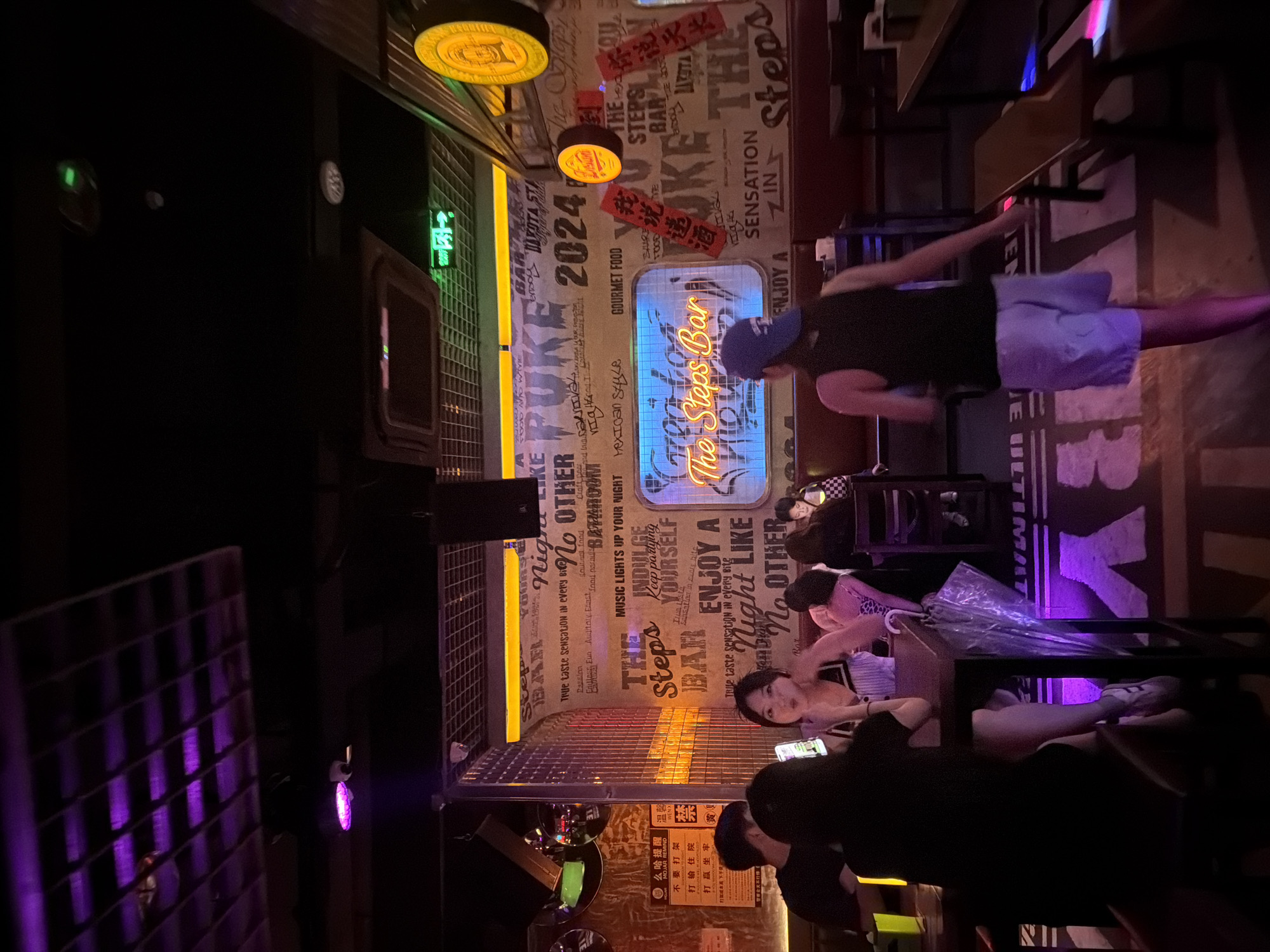 The interior of "The Steps Bar," located in Chongqing High-tech Zone, China, is captured at night. The walls are adorned with neon signs and text in both English and Chinese. Patrons are seated at tables, some looking at their phones. A man in a black tank top and blue shorts is walking through the space. The lighting is a mix of neon and purple hues.
