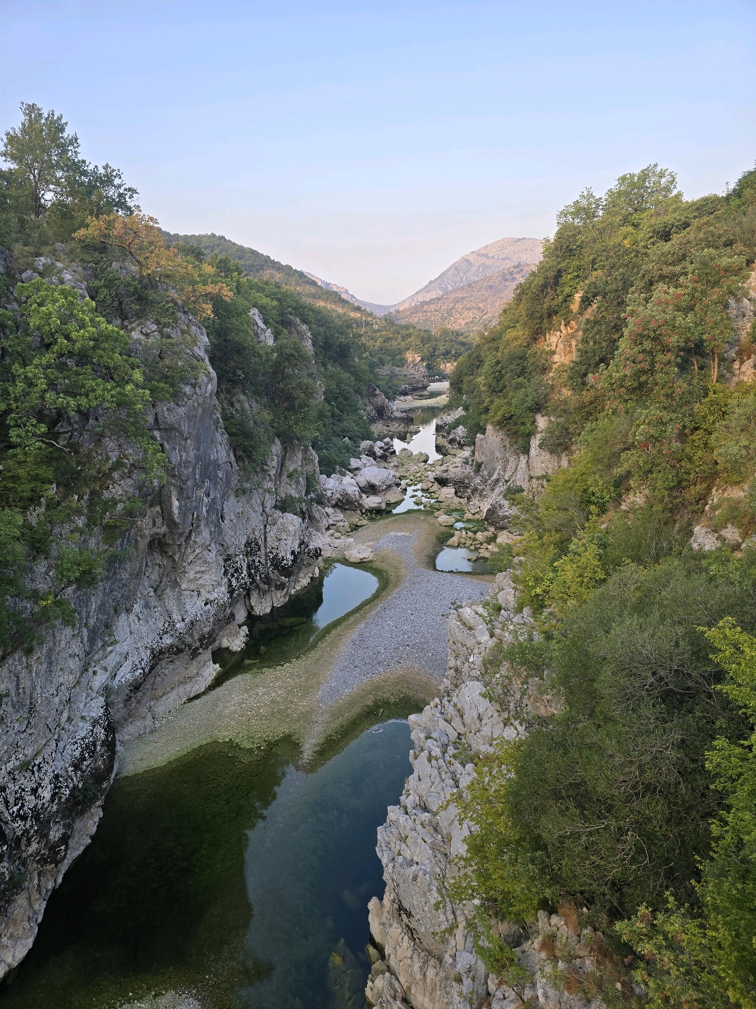 This is a high-angle, wide shot of a river canyon near Bioče, Montenegro. The canyon walls are steep and rocky, covered in lush green vegetation and trees. The river itself is shallow in parts, with exposed rocky riverbeds and small pools of dark green water. In the distance, more mountains are visible, fading into a hazy sky. The light suggests it is either early morning or late afternoon. There are no people or man-made structures visible in the image.