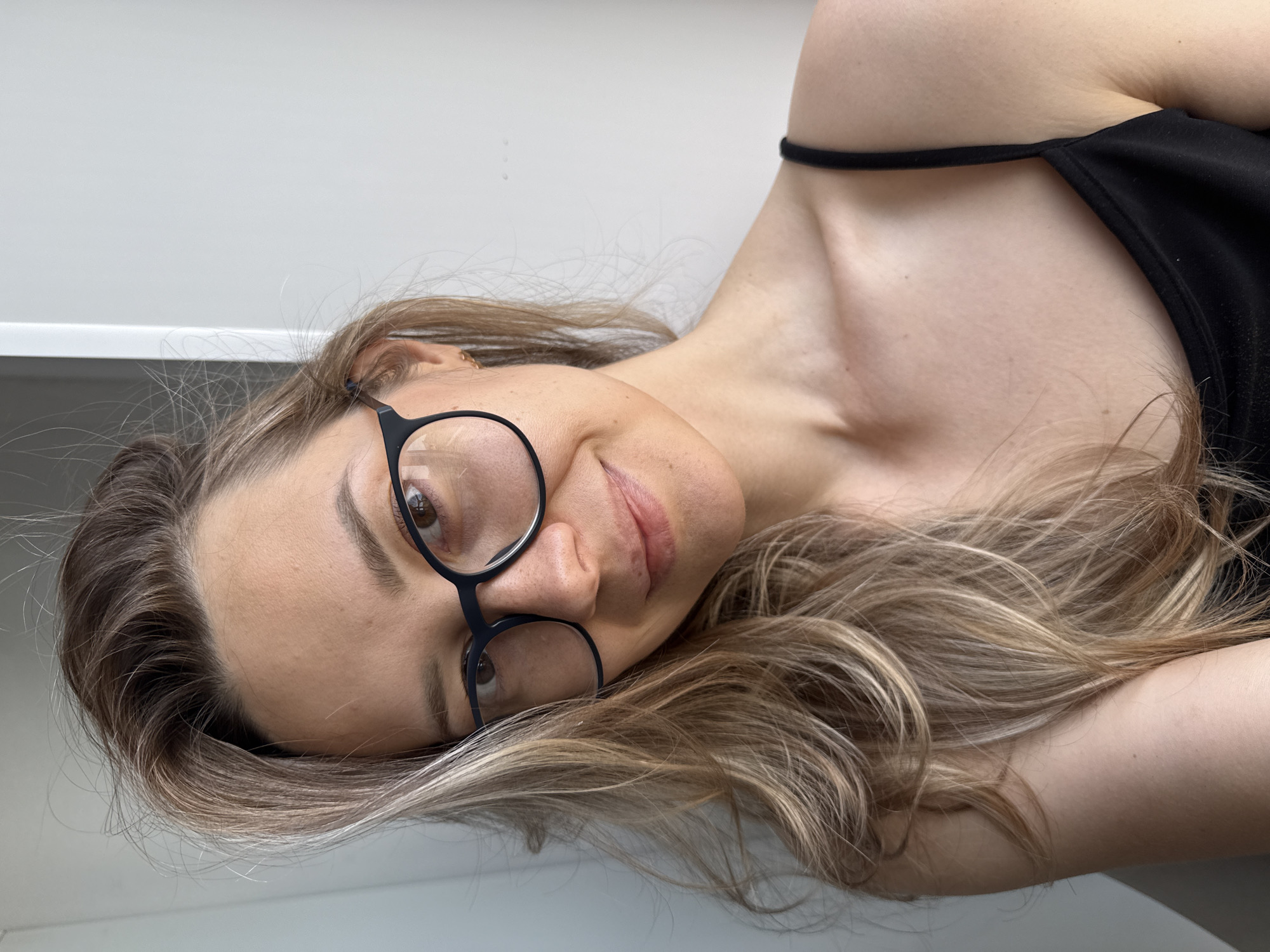 A portrait-oriented image, rotated 90 degrees clockwise, captures a young woman looking directly at the camera, likely in Chaikovsky, Russia. The woman has long, wavy hair, a blend of light brown and blonde with noticeable highlights, which is spread around her as she lies down. She is wearing black-rimmed eyeglasses, behind which her brown eyes are visible. Her expression conveys a calm and relaxed demeanor, with a subtle, gentle smile playing on her lips. She is dressed in a simple black spaghetti strap top. The background is a plain, light-colored surface, possibly a wall or a headboard, suggesting an indoor setting with soft, even lighting. Her skin appears fair. No visible text is present in the image.