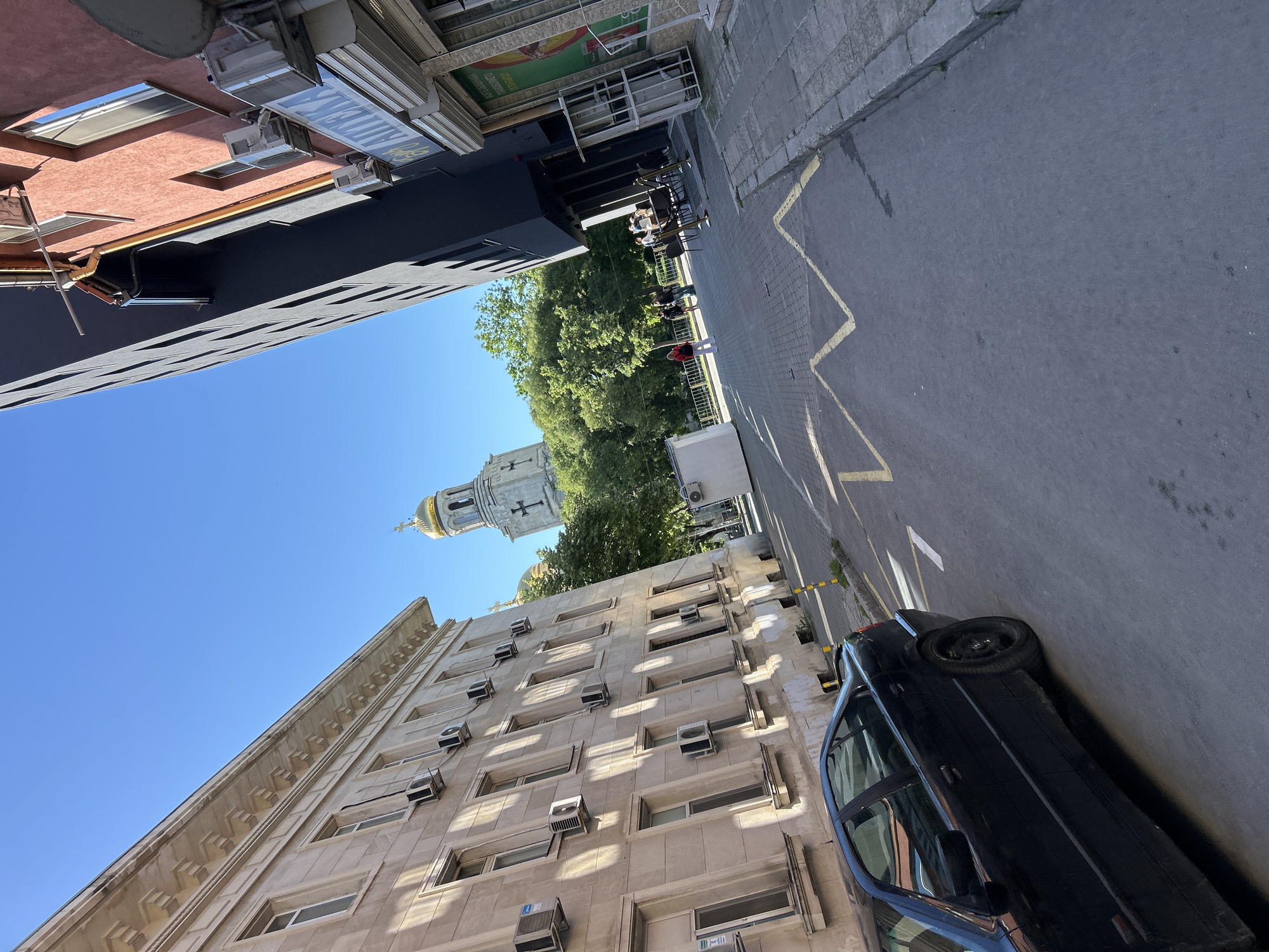 Captured from a ground-level perspective, the scene unfolds within the urban environment of Varna, Bulgaria. The vista includes sections of city streets and buildings, crowned by a view of the sky. A black car is parked on the roadside alongside a light-colored, multi-story building equipped with numerous window-mounted air conditioning units. A street runs adjacent to the building. The street is marked with yellow lines. In the background, between buildings, trees are visible along with a church with a distinctive golden dome. Along the street in the background, a few people are walking. Adjacent to the street a restaurant is seen with customers sitting at tables.