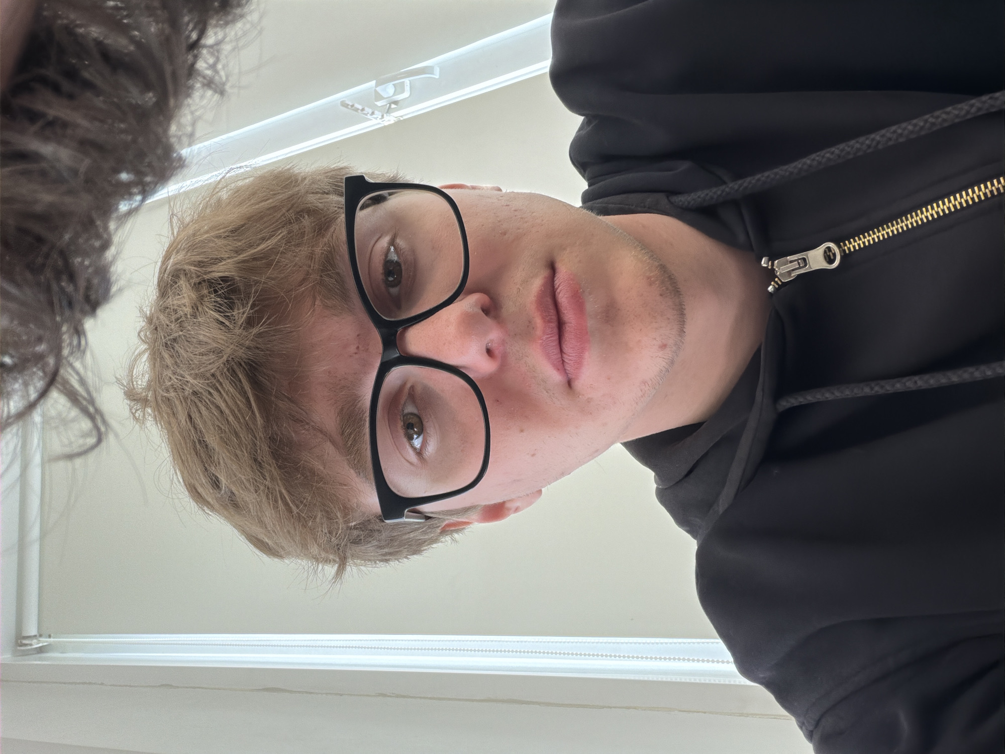 A close-up, downward-angled photograph features a young man with short, blond hair and glasses wearing a black hooded sweatshirt. He is positioned in front of a window with white blinds. The photograph also captures part of another person's hair on the left side of the frame. There is no information to verify that the picture was taken in Krasnoyarsk, Russia.