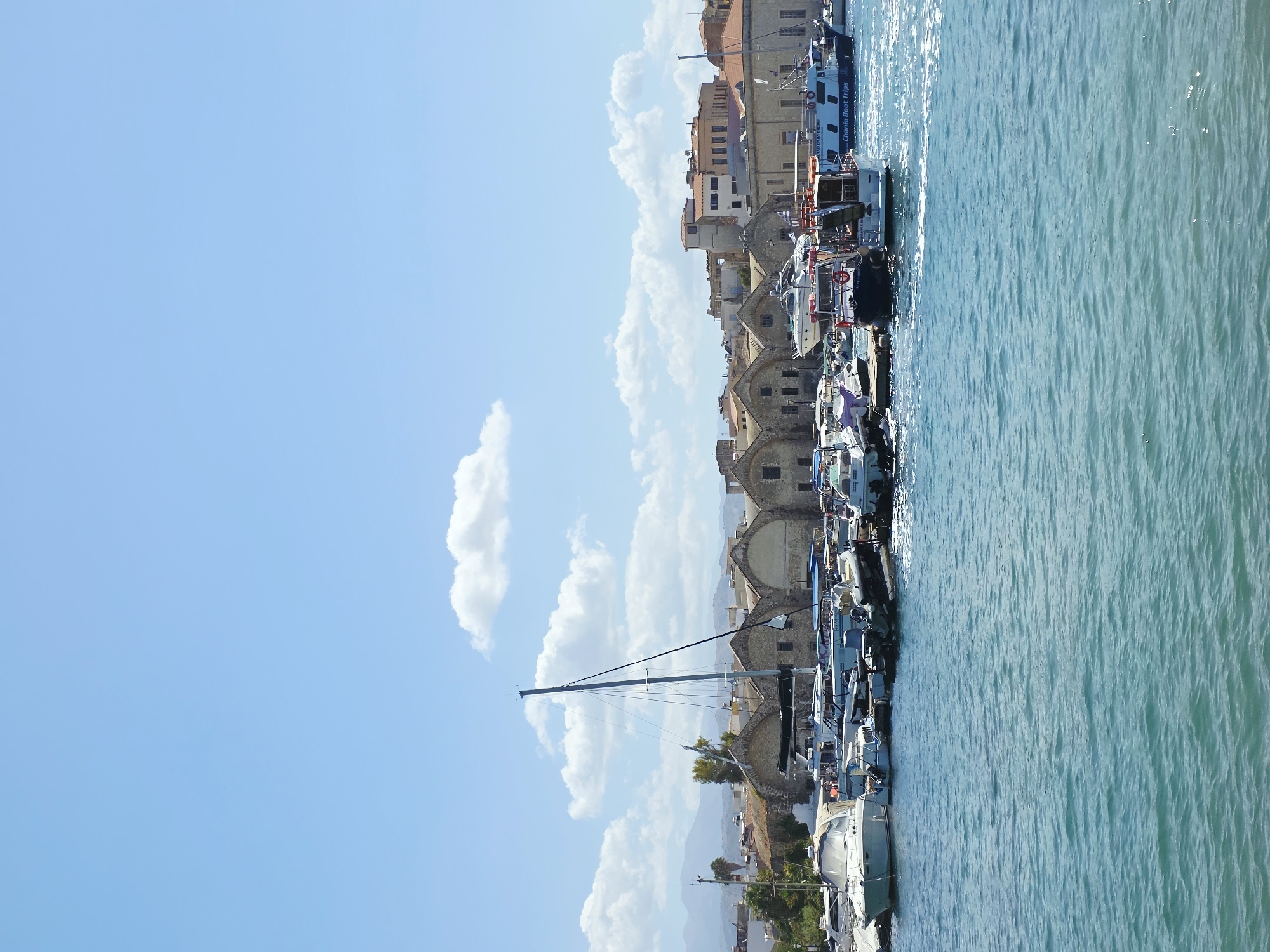The image captures a picturesque view of the Venetian Harbor in Chania, Greece. In the foreground, the turquoise waters of the harbor are visible, with gentle ripples reflecting the sunlight. A variety of boats are moored along the waterfront, ranging from sleek white motorboats to a classic sailboat with its mast standing tall.
In the middle ground, a row of historic Venetian-era buildings lines the harbor. These structures, characterized by their stone facades and arched openings, appear to be former warehouses or arsenals, showcasing the rich history of the port. Some of these buildings are topped with red-tiled roofs, adding a splash of color to the scene. A few palm trees are interspersed among the buildings, hinting at the Mediterranean climate.
Further in the background, the landscape transitions to a hazy mountain range, likely part of the White Mountains of Crete, under a bright blue sky dotted with fluffy white clouds. The overall atmosphere is serene and idyllic, suggesting a pleasant day in this charming Greek city. The scene is devoid of any prominent human activity, emphasizing the architectural beauty and the tranquil maritime environment.