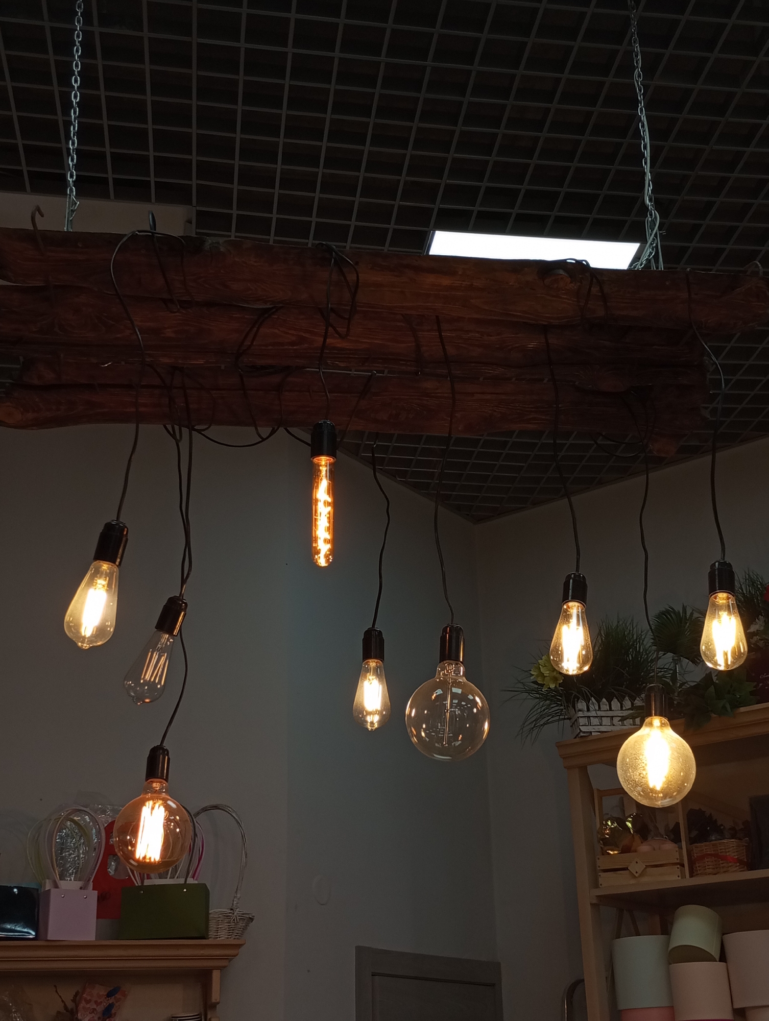 The image depicts a lighting fixture consisting of a rough-hewn wooden beam suspended from the ceiling by chains. Several Edison-style light bulbs of varying shapes and sizes hang from the beam by black wires. The ceiling is a dark grid pattern, with a bright white rectangular light fixture visible in the background. Below the hanging lights, shelves display various decorative items, including plant pots and figurines. The scene appears to be indoors. While the location is not definitively identifiable from the image, the question context indicates it is Izhevsk, Russia. No people are visible in the image.