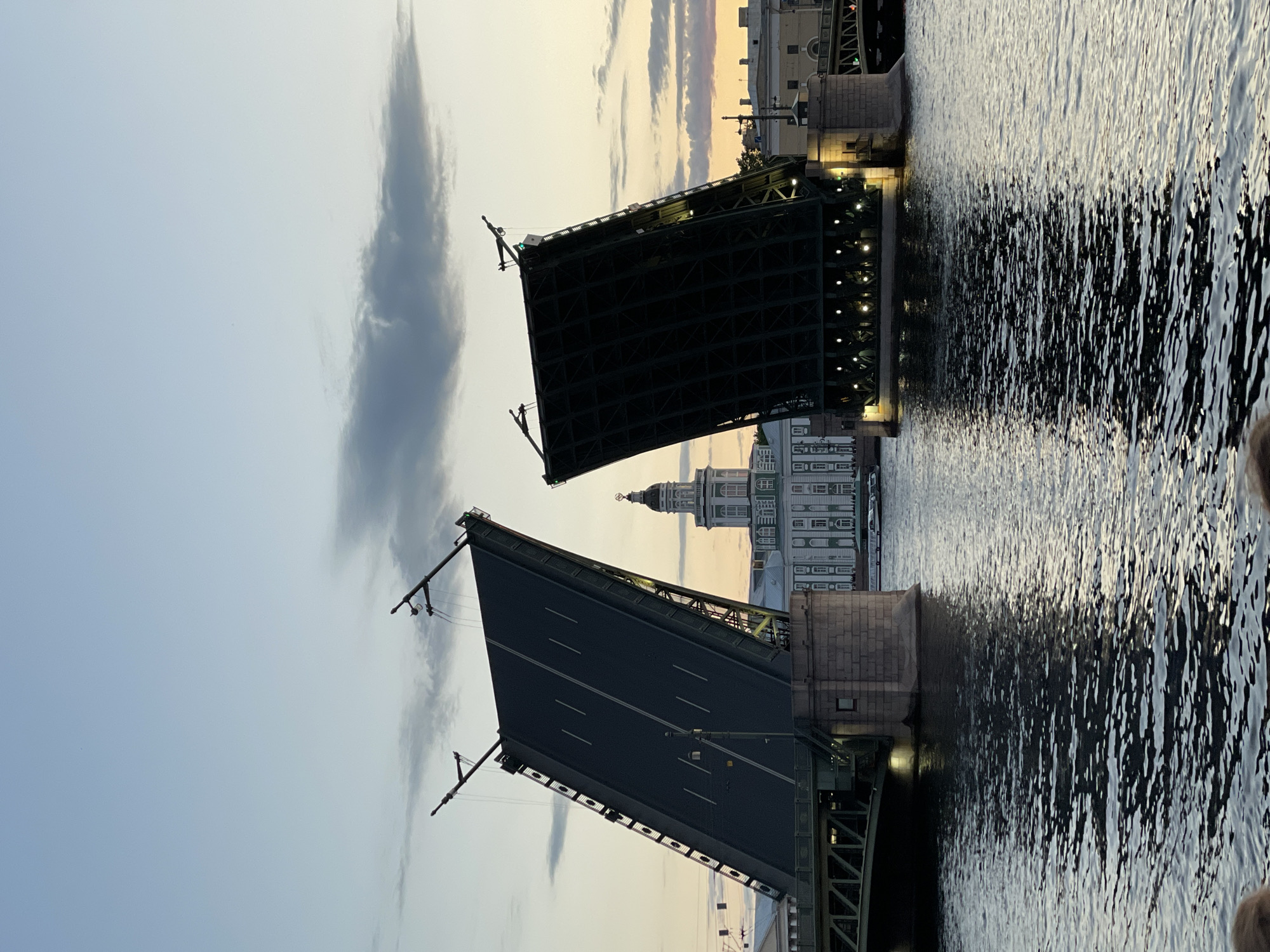 In an outdoor, waterfront scene, a bascule bridge is raised, with its two halves angled upwards in the sky. The bridge spans a body of water, likely a river or canal, with gentle ripples on the surface. The sky above is a gradient of pale blue near the horizon, transitioning to a soft grey with wispy clouds higher up. The scene appears to be set during dusk or early dawn, as suggested by the warm, diffused light.
In the background, a large, light-colored building with a central dome and windows is visible. Its architecture suggests a historical or institutional purpose. Further in the distance, other buildings and structures form a cityscape, with some lights illuminating the facades. The bridge itself is a dark color, possibly green or black, and is illuminated by strings of lights underneath its raised sections. The stonework of the bridge's supports is visible, adding a sense of solidity to the structure.
There are no people clearly visible in the foreground or on the bridge. The overall impression is one of architectural grandeur and urban infrastructure set against a serene natural backdrop.
