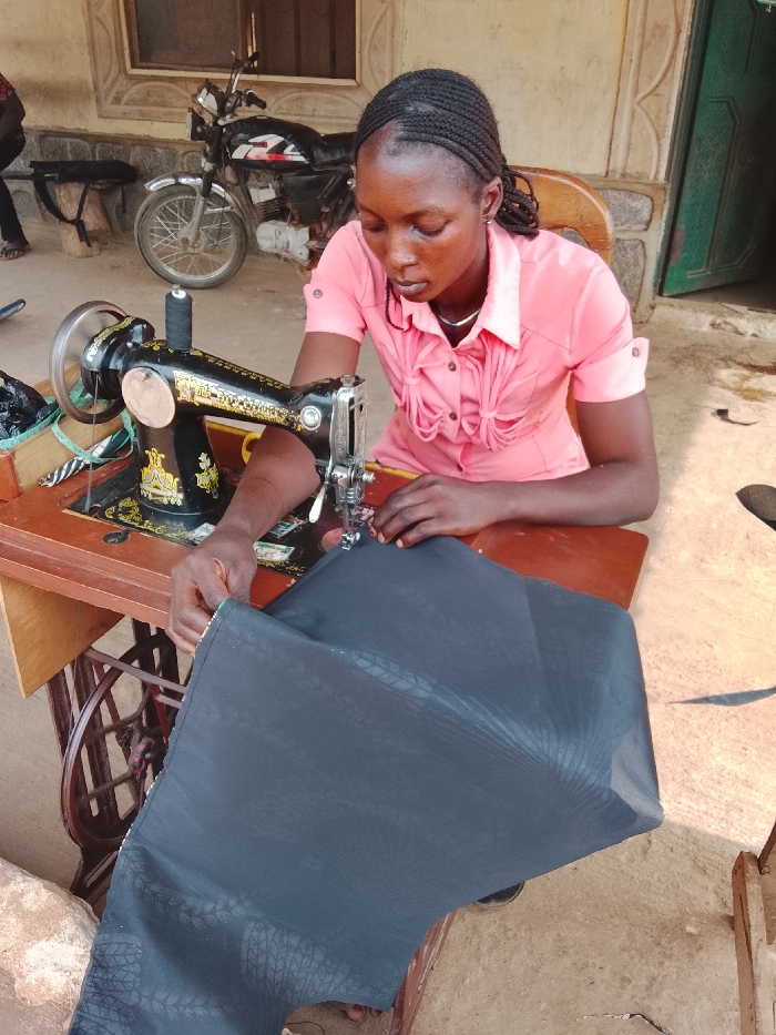 In Akwanga, Nigeria, a young individual, identified as female, with braided dark hair, is seated and engaged in tailoring activities. She wears a short-sleeved coral-pink collared shirt and is intently operating a black vintage-style sewing machine, characterized by gold decorative accents. Her hands guide a piece of dark grey fabric, featuring a subtle textured pattern, under the machine's needle. A spool of black thread is present on the machine's spindle. The sewing machine rests on a wooden table, supported by a metal treadle stand, parts of which are visible.
The scene unfolds outdoors or in an open-air workshop. In the background, to the left, a black motorcycle with chrome components is parked. Behind the motorcycle, a light-colored building wall with molded architectural details is visible, along with the bare feet of another person. To the right of the individual, a wooden chair is positioned, and a dark-framed doorway leads into an interior space. The ground surface is uneven, light-colored, and appears to be concrete or packed earth, illuminated by natural daylight.