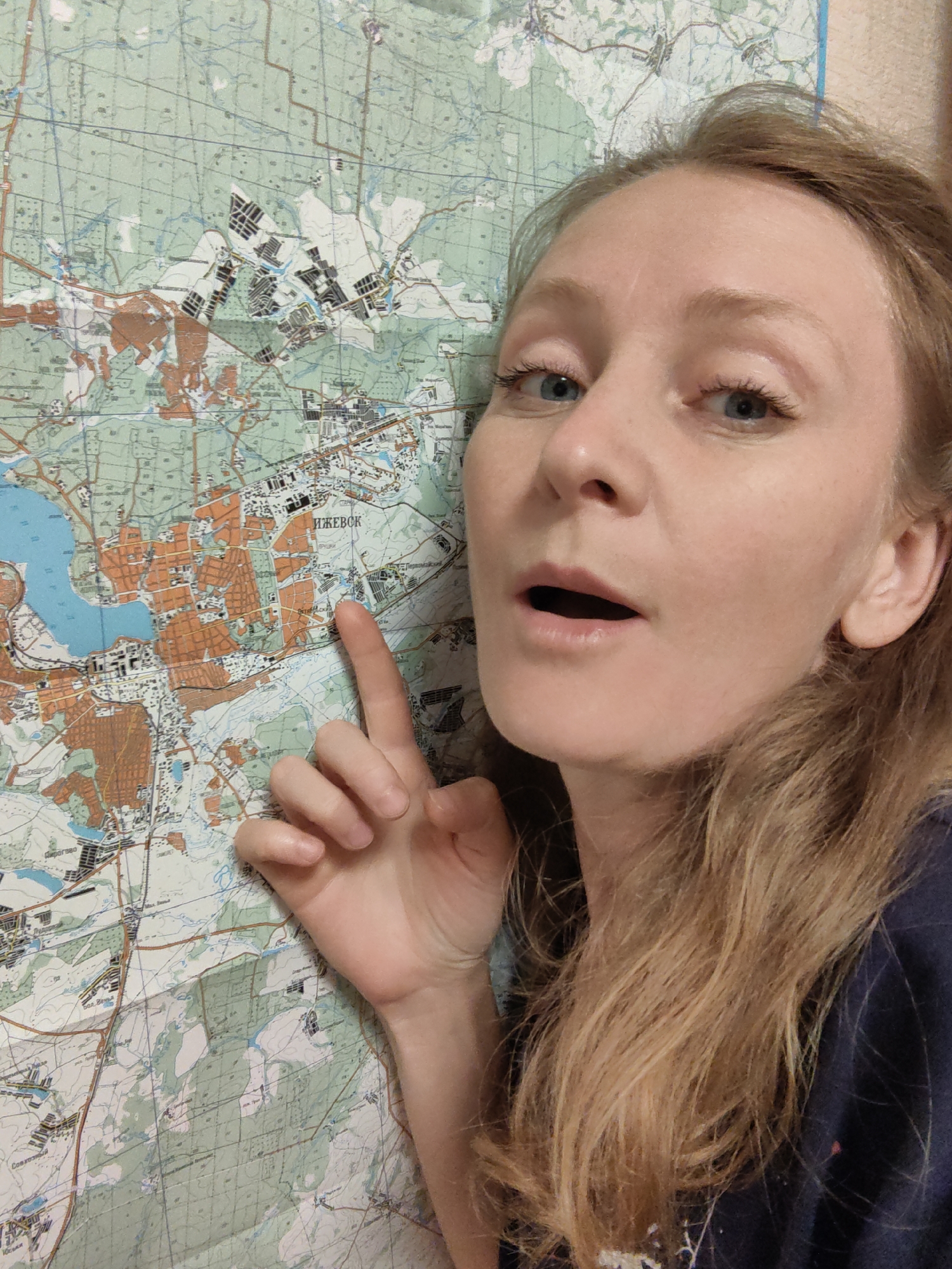 A woman points with her right index finger at a large, folded topographical map, positioned on a wall behind her. The map prominently displays the city of Izhevsk, Russia, with the name "ИЖЕВСК" (IZHEVSK) clearly labeled in Cyrillic letters. The woman's finger is pointing towards an area within the city's urban layout. The map appears to denote various landscape features. The woman has fair skin, blue eyes, and light brown hair and is wearing a dark shirt. Her mouth is open in what appears to be a gesture of surprise or announcement. The map colors are predominantly green, brown, and blue.