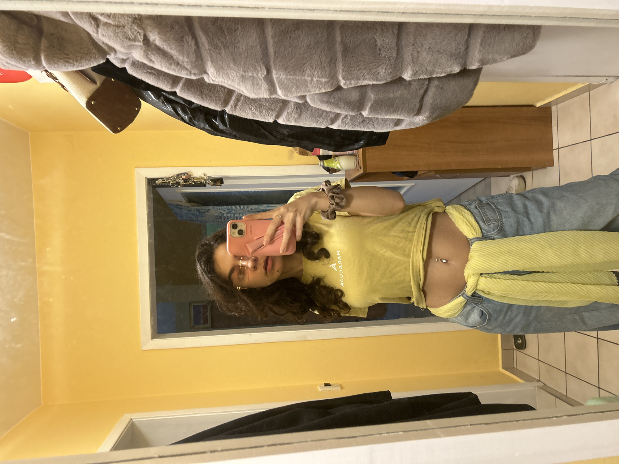 The image is a selfie taken in front of a mirror. The subject is a young woman with curly dark hair, wearing gold-rimmed glasses, a yellow t-shirt knotted at the waist, light blue jeans, and a yellow scarf tied around her waist. She is holding a pink iPhone in her right hand, taking the picture. The room has yellow walls and white trim. In the mirror's reflection, part of a room with a blue bedspread and framed artwork can be seen. Hanging above the mirror on the left is a garment bag, and above that is a grey fluffy coat. A brown wooden cabinet is visible on the right side of the mirror, along with white tile flooring. The location, Sofia, Bulgaria, cannot be determined from the details in the image.