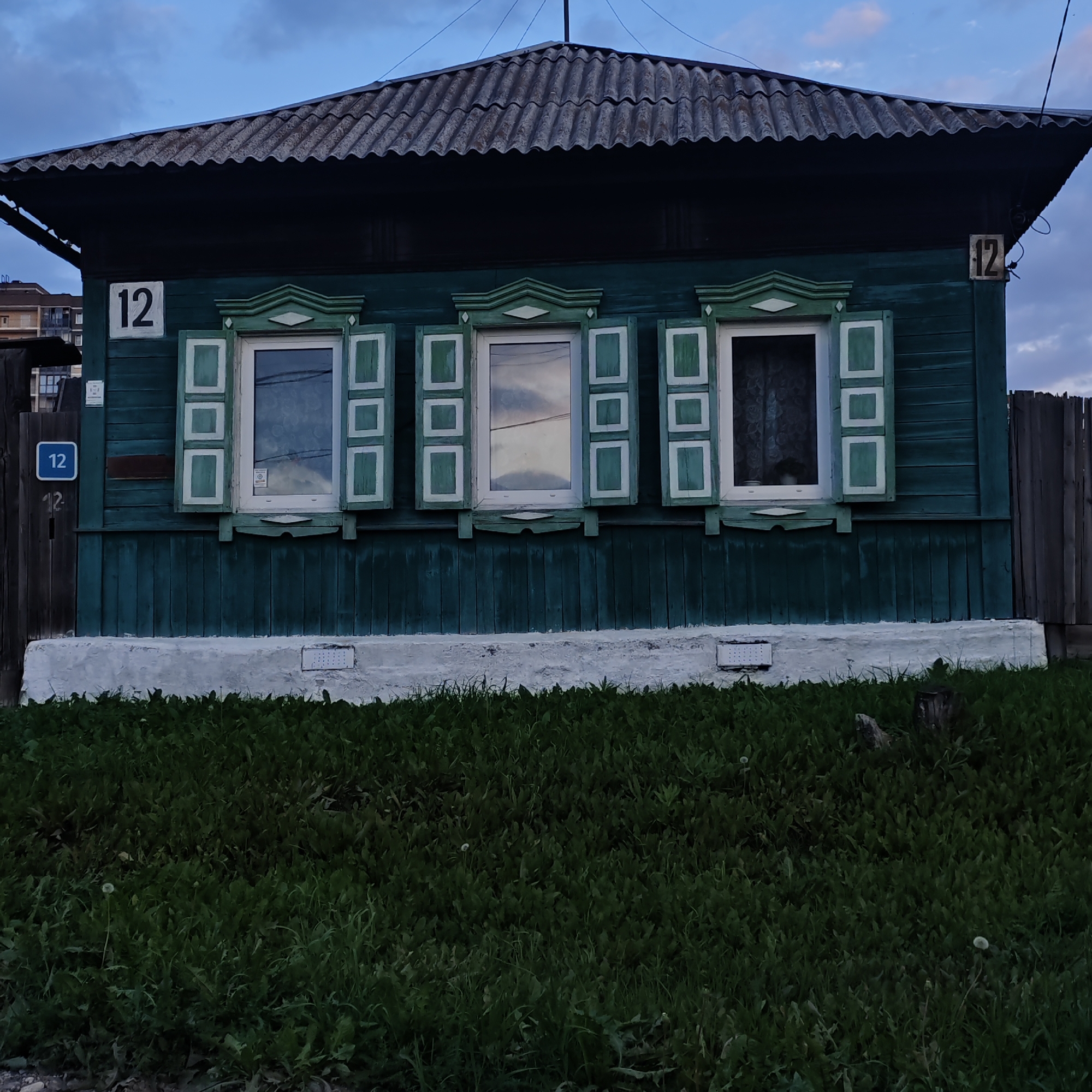 The image shows the facade of a dark green wooden house with three windows featuring decorative shutters. The house has a corrugated roof and a white foundation. A wooden fence runs along the right side of the house. A wooden post with a blue sign displaying the number "12" is visible to the left of the house. Another sign with "12" is mounted on the house to the left of the windows. The foreground is covered with green grass and dandelions. The sky is cloudy and appears to be dusk. This appears to be a residential property in Irkutsk, Russia, likely in a less developed area given the architectural style of the house and the presence of a wooden fence. There are no people visible in the image.
