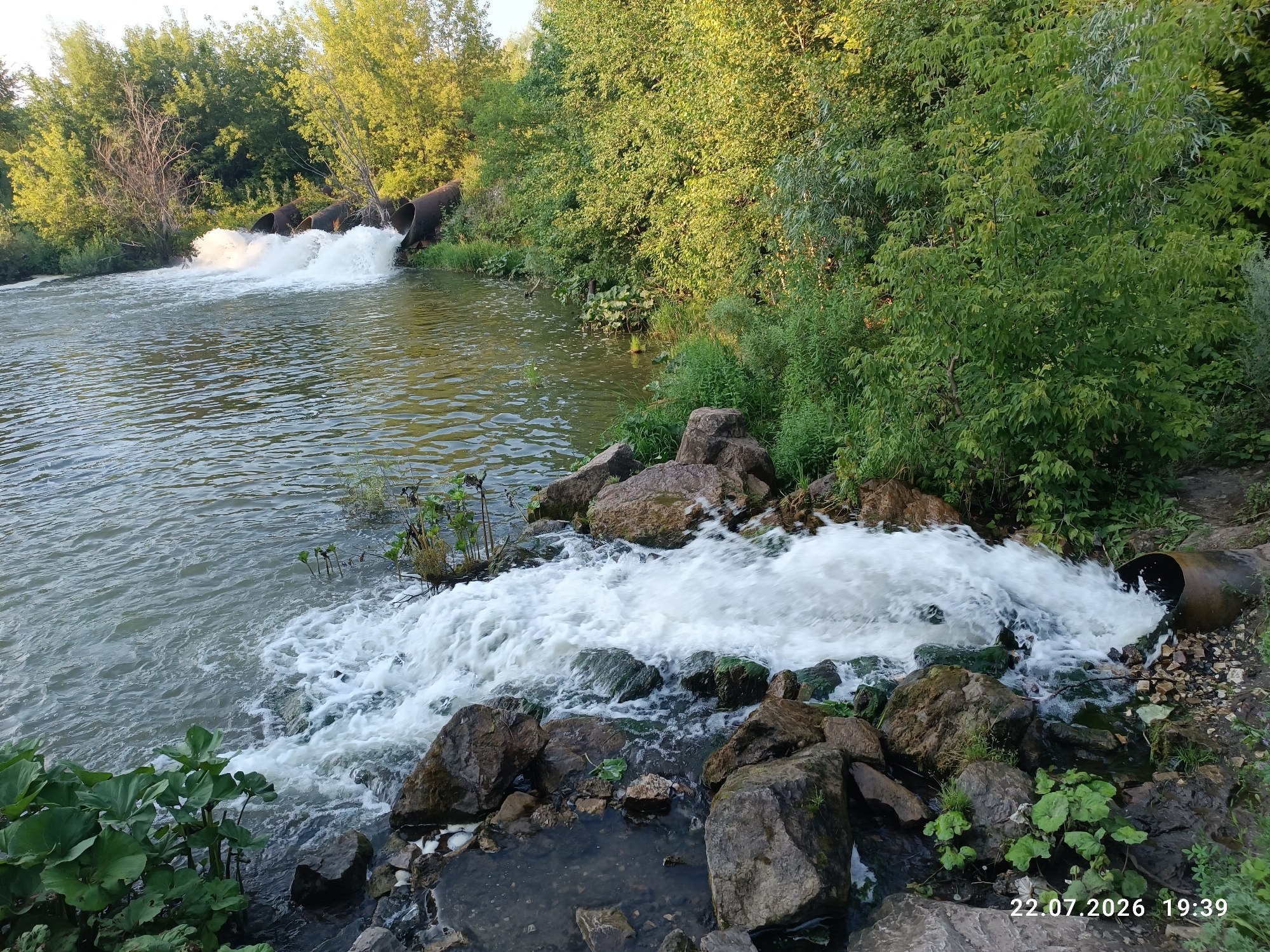The image depicts a body of water with multiple large pipes discharging a significant volume of foamy white water. These pipes are situated along a bank lined with dense green vegetation and trees. The water surface shows ripples, indicating movement. In the foreground, several large, dark rocks are visible, some of which are partially submerged in the water. The scene is bathed in a warm, natural light, suggesting it is either early morning or late afternoon. A timestamp in the bottom right corner reads "22.07.2026 19:39". The location is identified as Verkhotor, Russia. There are no people or animals visible in the image.