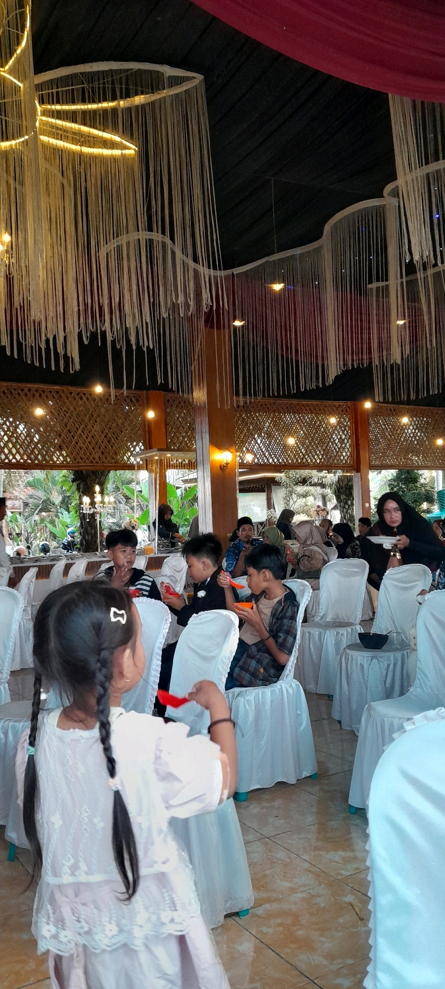 The image depicts an indoor gathering, possibly a wedding reception or a banquet, in Padang, Indonesia. The venue appears to be decorated with numerous hanging white cords and strings, some illuminated with yellow lights, creating a decorative ceiling feature. Wooden beams and lattice work are visible, suggesting an open-air or semi-open-air setting with natural elements. Several individuals are seated at tables, covered with white cloths. In the foreground, a young girl with braided hair and a white dress is holding a red spoon, facing away from the camera. Several boys are seated at tables, eating. One boy in a black suit is holding an orange spoon, while another boy in a plaid shirt is also eating. In the background, other guests are visible, including a woman in a black hijab holding a plate. The flooring is tiled. The overall atmosphere appears to be festive and communal.