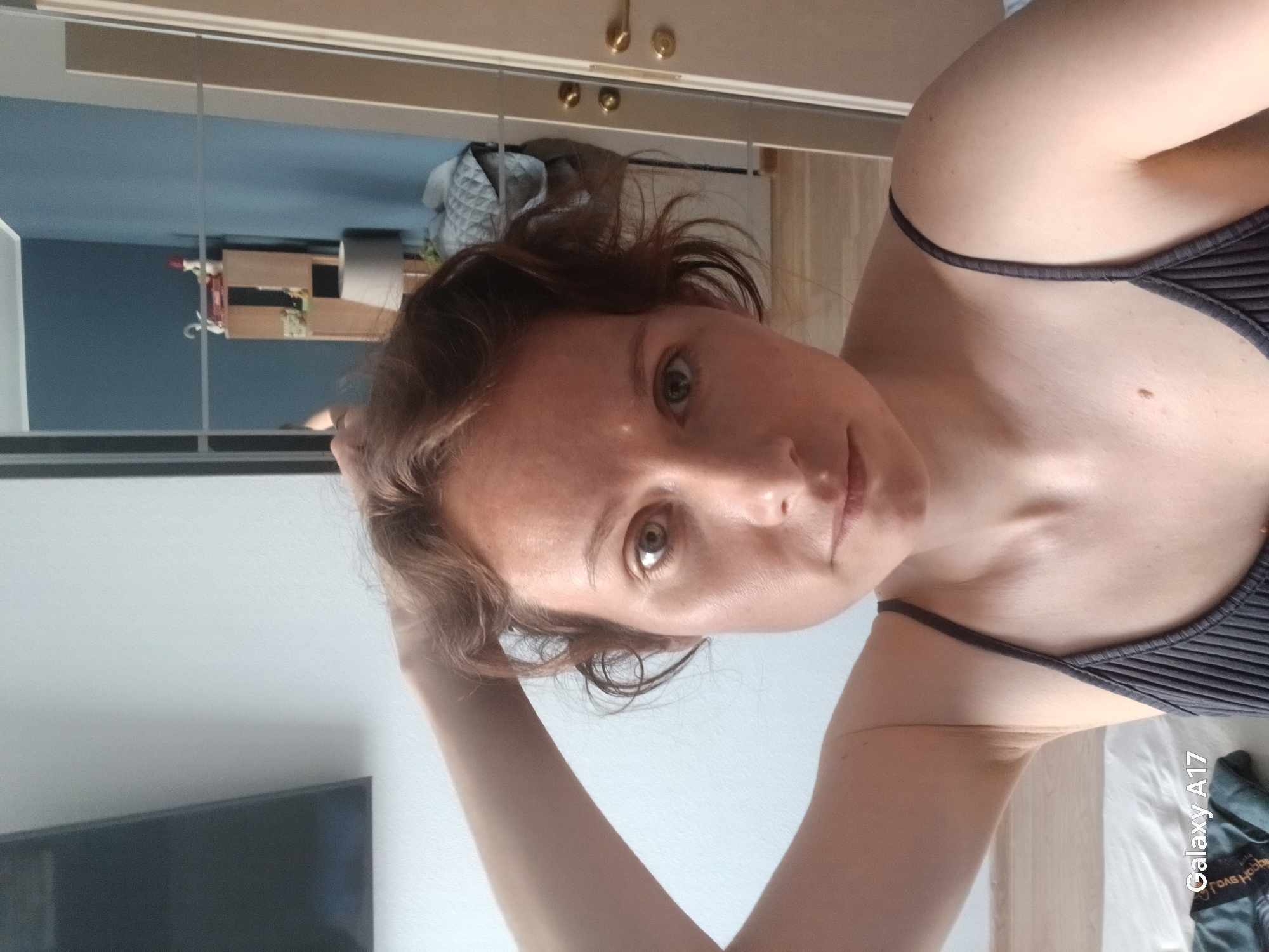 A woman with light skin and brown hair is posing for a selfie. She is wearing a dark-colored top with thin straps. Her left hand is near her head. The background appears to be a room with mirrored closet doors, reflecting part of the room. There is a television visible in the bottom left corner of the image, and furniture is visible in the reflection. The photo was taken with a Galaxy A17 phone. The image provides no information regarding the city of Yekaterinburg, Russia.