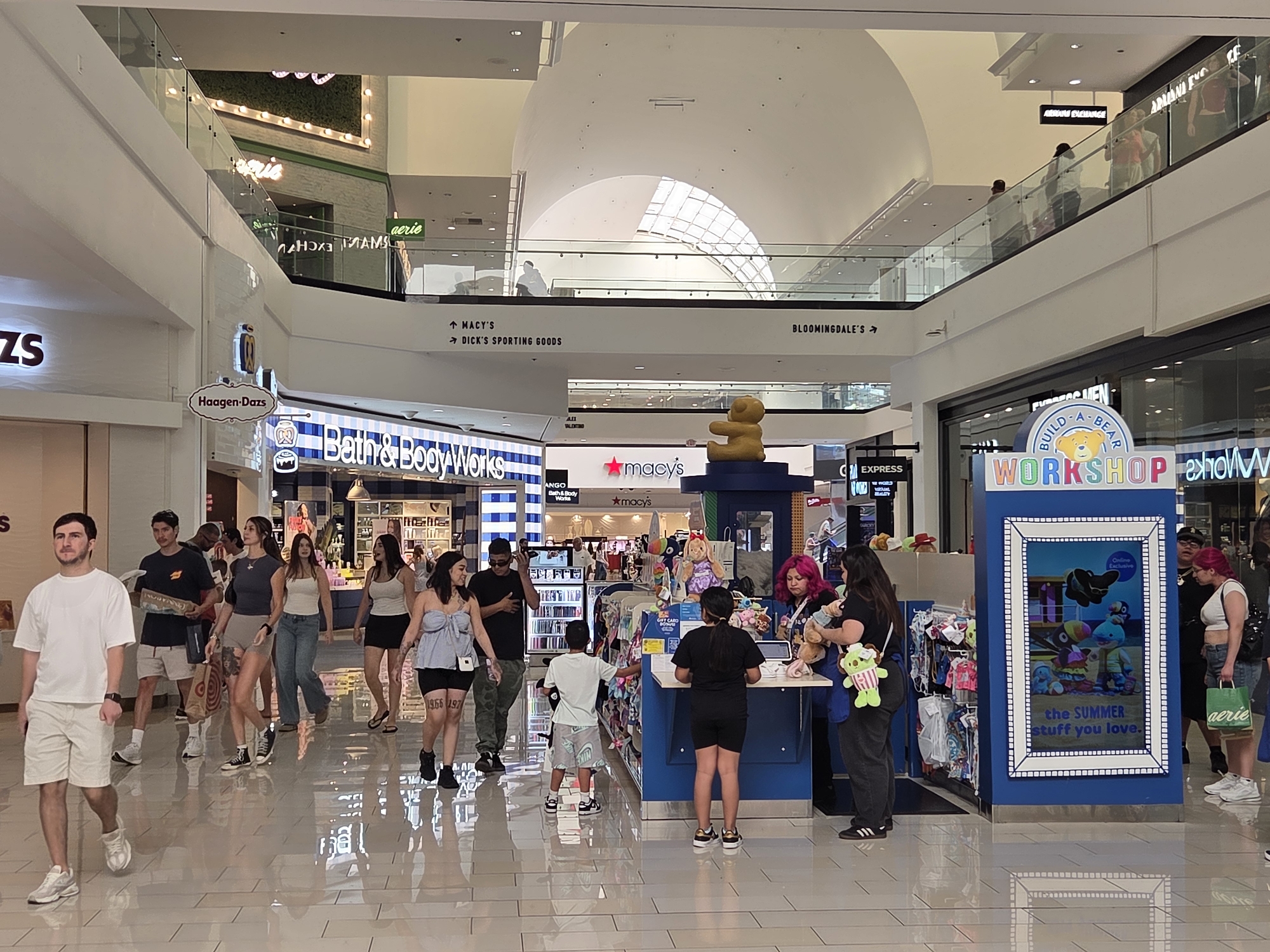 The image depicts a bustling interior view of the Glendale Galleria mall in Glendale, California. The scene is a wide shot looking down a brightly lit, polished tile hallway. Several people are walking and interacting within the mall's common area.
On the left side of the frame, a young man in a white t-shirt and khaki shorts walks towards the camera. Further back, a group of people are seen walking, some carrying shopping bags. Signs for stores like "Haagen-Dazs" and "Bath & Body Works" are visible, indicating the retail environment.
In the center of the image, a vibrant blue "Build-A-Bear Workshop" booth is prominently displayed, featuring colorful advertisements and plush toys. A few individuals are interacting with the booth and staff, including a child standing at the counter. The background shows more stores and the upper levels of the mall with people browsing.
The architectural style is modern, with glass railings on elevated walkways and a high, domed ceiling that allows natural light to filter in. The overall atmosphere suggests a typical busy shopping day at a large indoor mall.