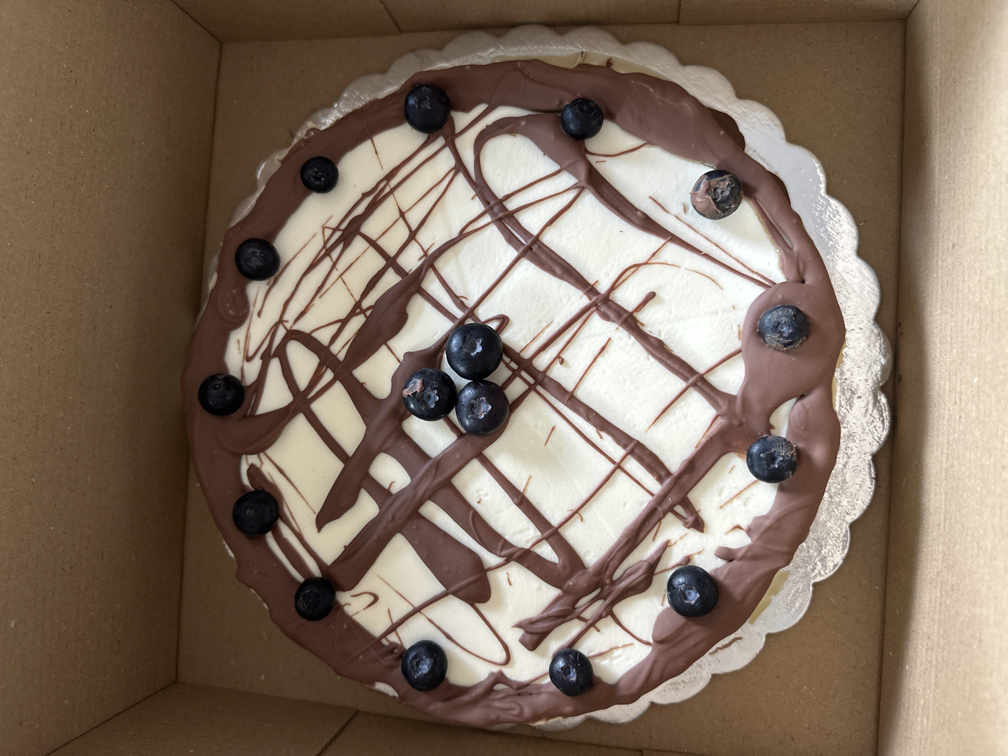 A whole round cake sits inside a cardboard box. The cake has a white frosted top drizzled with lines of brown chocolate. Blueberries are arranged around the edge of the cake and in a small cluster in the center. The cake rests on a silver, scalloped-edged cake board. The box is plain, light brown cardboard. There is no indication of the location being Lehchevo, Bulgaria in the image.
