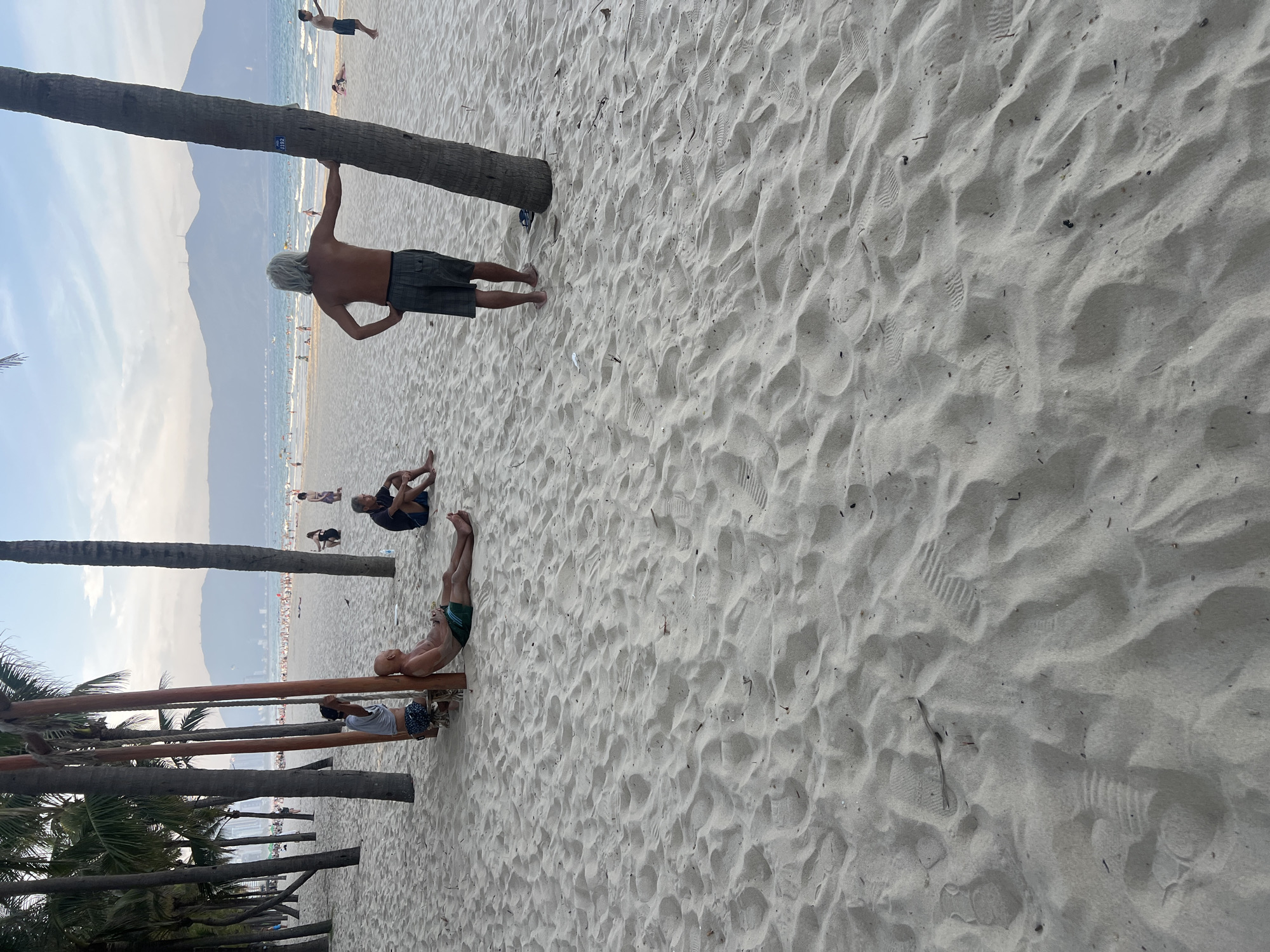 This image captures a section of My Khe Beach in Da Nang, Vietnam, under a hazy sky. The foreground is dominated by soft, pale sand, showing numerous footprints and some scattered debris. In the mid-ground, several people are present. To the left, three men are clustered near a wooden swing set with large palm trees. One man sits on the ground leaning against a wooden post, another sits on the swing, and a third stands leaning against a palm tree. Further to the right, a man with long, gray hair stands with his hand on a palm tree, wearing shorts. In the background, the ocean stretches out, with waves visible and several people swimming or sunbathing on the shore. A distant mountain range forms the horizon. The overall impression is a relaxed, casual beach scene with a mix of locals and possibly tourists enjoying the seaside.