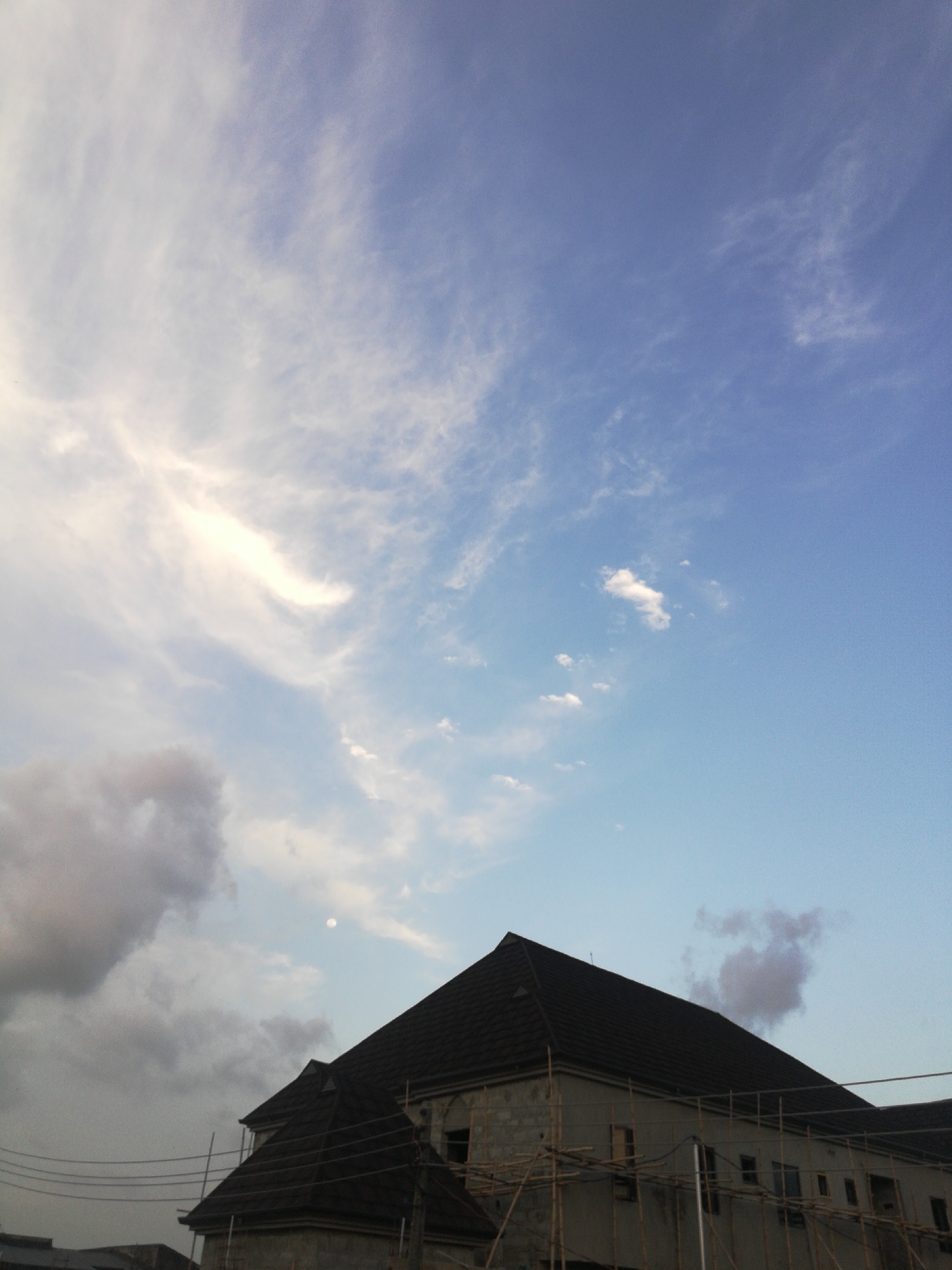 This media file captures a scene featuring a developing urban landscape under a dynamic sky in Lagos, Nigeria. The upper two-thirds of the image are dominated by the sky, which transitions from a brighter, lighter blue with wispy white clouds on the left to a deeper blue with more scattered, smaller white clouds on the right. A distinct, bright, spherical object, likely the moon, is visible in the upper right quadrant of the sky, indicating the photograph was taken during daylight hours or twilight.
In the lower portion of the image, a large, multi-story building is prominently featured. It has a dark, steeply pitched roof and light-colored walls, suggesting it's constructed from concrete blocks. The building is clearly under construction, as indicated by the extensive bamboo scaffolding erected around its exterior. Multiple rectangular window openings are visible on its facade.
To the far left and lower down, denser, darker grey clouds are present, contrasting with the lighter, clearer parts of the sky. Power lines are visible stretching across the lower left area, adding to the urban context. No people are visible, nor is any discernible text. The scene conveys a sense of ongoing development and construction in the region.