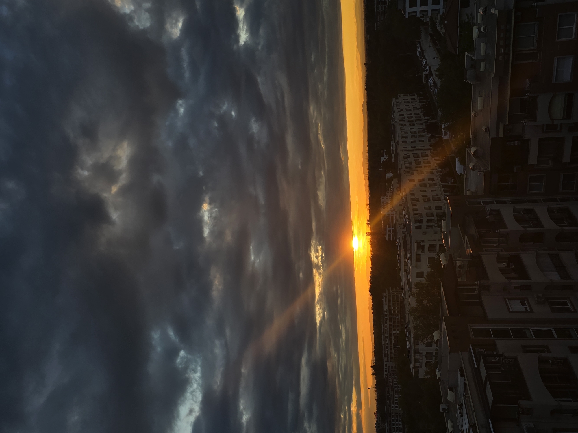The image shows a sunset in an urban setting. The sky is filled with dark, stormy clouds on the left, contrasting with a bright orange and yellow sunset on the right. The sun is positioned on the horizon, with beams of light shining through the clouds. Buildings and trees are visible along the horizon, silhouetted against the vibrant sky. The foreground features residential buildings, suggesting the location is a city.