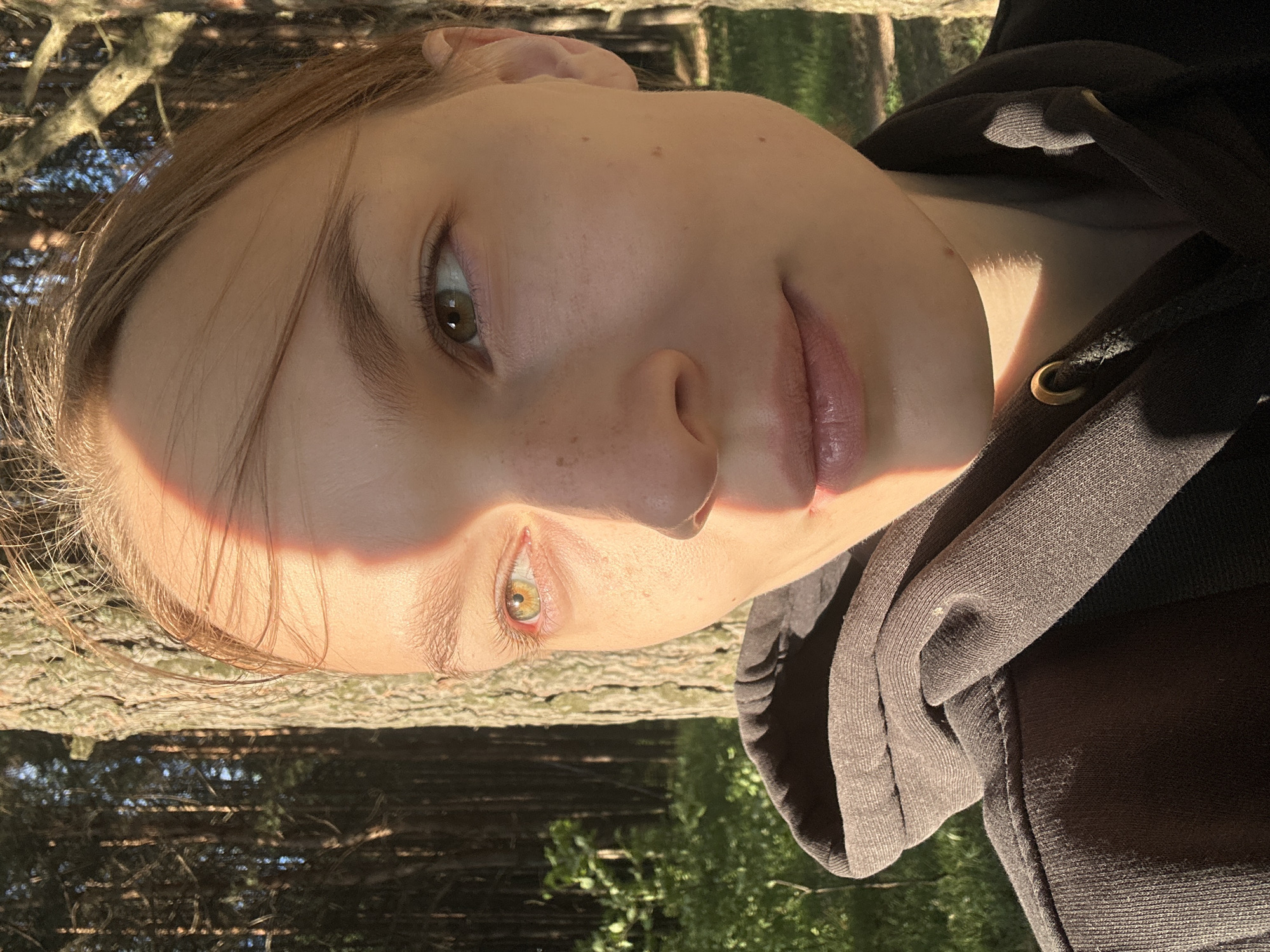 This is a close-up, portrait-style photograph of a young person with light brown hair. The subject is looking upwards and to the left, with their head tilted. Sunlight is casting a diagonal stripe across their face, highlighting their right eye and cheekbone. They have light skin with a few freckles visible. Their eyes are a hazel or light brown color, and they are wearing light pink eyeshadow. Their lips are slightly parted and appear to be a natural pinkish-red color.
The subject is wearing a dark grey hooded sweatshirt. The texture of the fabric is visible, including some subtle pilling. The drawstring for the hood is visible, with a metal grommet.
The background is blurred, suggesting an outdoor setting. There are hints of tree bark and foliage, indicating they are likely in a wooded area. The lighting suggests it is daytime, possibly late afternoon or early morning, due to the warm, angled sunlight. The overall mood of the image is serene and natural. No text is visible in the image.
