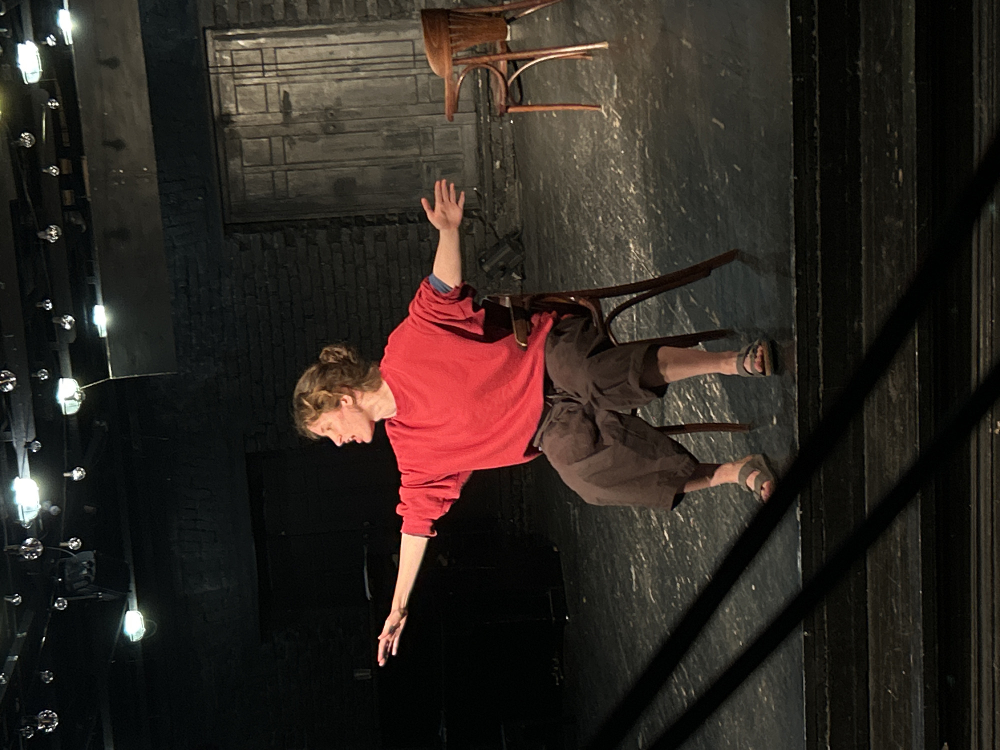 On a dark stage with brick walls and string lights, a person sits on a wooden chair, wearing a red shirt and brown shorts. One arm is raised upwards while the other is extended downwards. Another wooden chair is precariously balanced above. The stage also features tiered steps. The location is Unknown, Unknown.