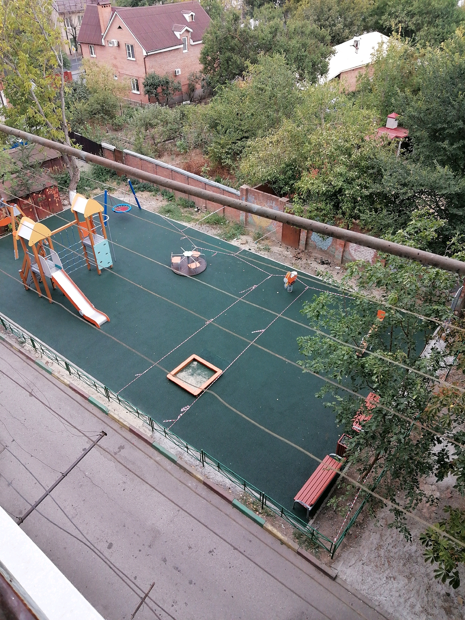A high-angle, overcast, outdoor photograph depicts a children's playground area adjacent to a street in Rostov-on-Don, Russia. The playground surface is a dark green, rubberized material, with red and white caution tape demarcating certain zones. Playground equipment includes a multi-level climbing structure with an orange and white slide, a circular merry-go-round, a spring rider in an orange and white color scheme, and a red-rimmed sandbox with a metal frame. Two wooden park benches with orange seating are situated near the edge of the playground, under the shade of leafy trees. The playground is bordered by a low green metal fence and a sidewalk with a red and yellow curb. A paved street runs alongside the sidewalk, with power lines suspended above it. In the background, a red-brick house with a dark red roof and multiple windows is visible amongst lush green trees and foliage. Another building with a white roof and chimney is partially obscured by trees. The overall impression is of an urban residential area with a dedicated play space for children.