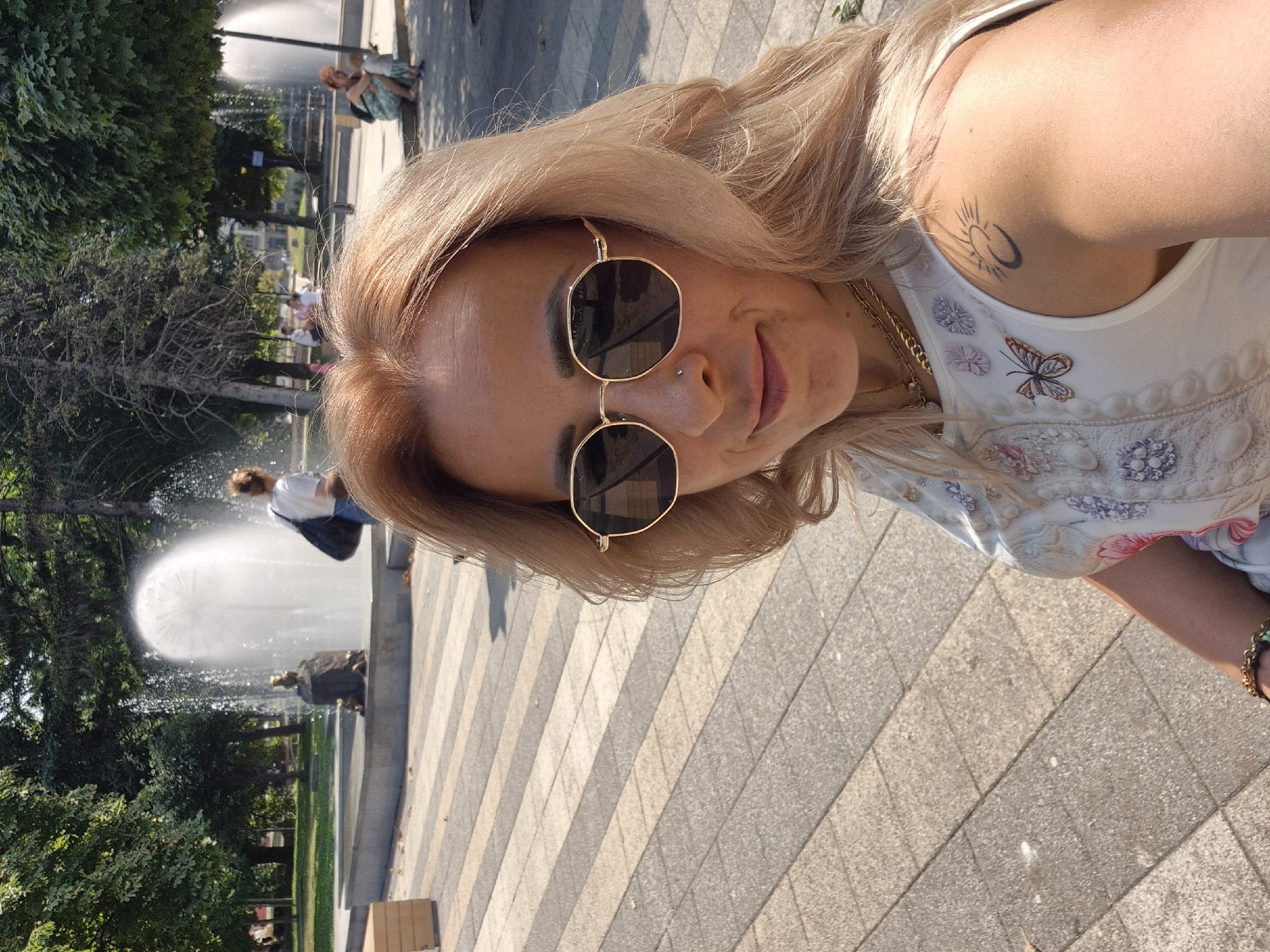 A woman with blonde hair and sunglasses is taking a selfie in a park. She is wearing a white top with embroidered flowers and a butterfly. She has a tattoo of a sun and moon on her right shoulder. In the background, there are fountains and trees. The ground is paved with geometric patterns.
