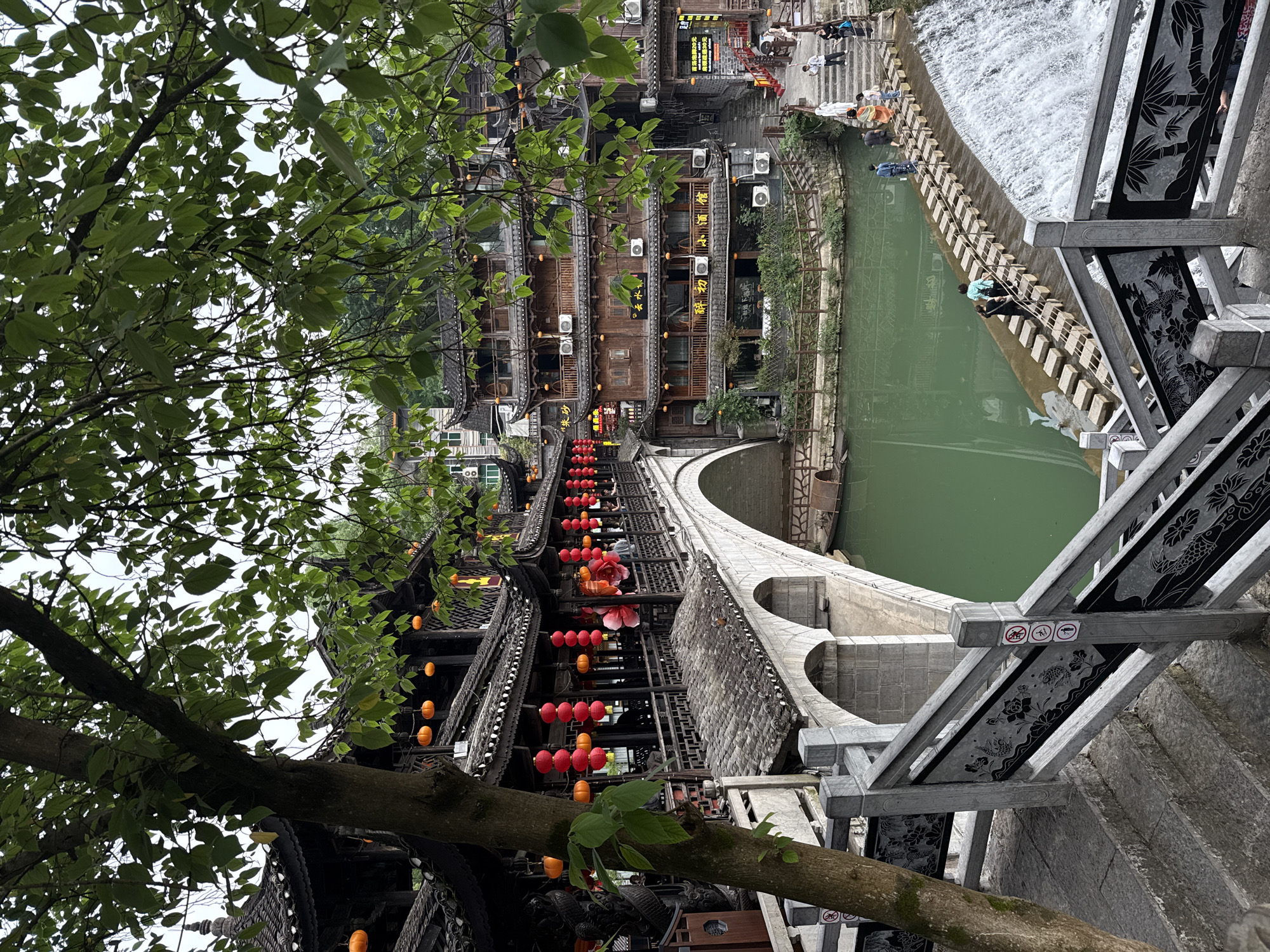View of Furong Ancient Town, Xiangxi, China. A stone arch bridge spans a green river with a waterfall at its end. Structures line the river, decorated with red and orange lanterns. People walk on the street in the distance. A tree frames the view on the left. Gray stone bridge railings with designs are visible on the right.