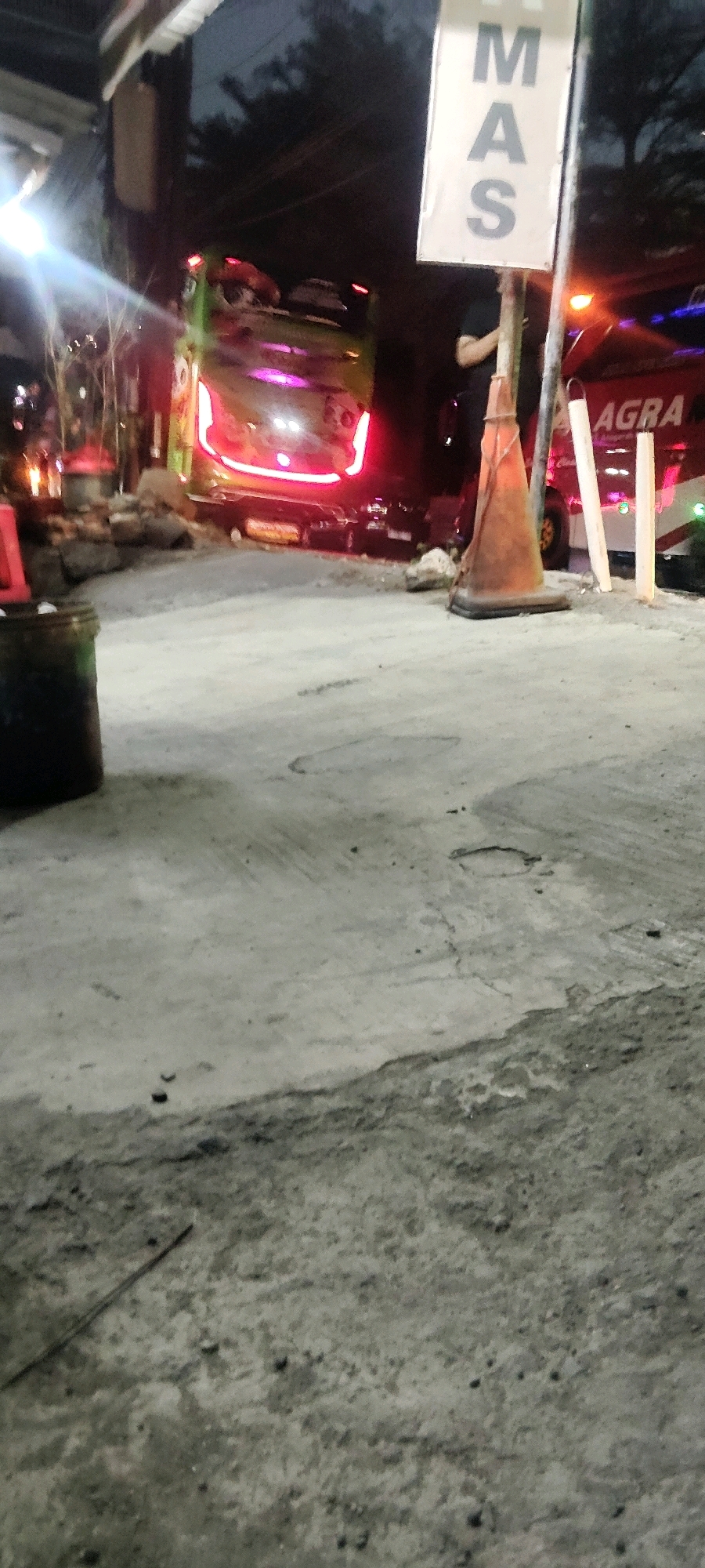 In the city of Pasirsari, Indonesia, a scene unfolds at what appears to be a roadside or transportation hub at dusk or night. A large, green and yellow bus with bright red taillights is prominently featured, facing away from the camera and partially obscured by debris or rocks. To the right of the bus, a red vehicle with the word "AGRA" visible on its side is parked.
In the foreground, a rough, grey concrete surface dominates, with visible cracks and imperfections. A dark, cylindrical object, possibly a bucket or trash can, is positioned on the left side of the image. A bright orange traffic cone is placed near the right side of the bus, its base resting on the concrete. Behind the cone, two vertical white posts stand.
A sign with the letters "MAS" in large, dark text on a white background is mounted on a pole near the center-right of the image. A person, dressed in dark clothing, is partially visible behind the sign, appearing to interact with or stand near the traffic cone. The background is dark and indistinct, suggesting trees or other structures, with some ambient lighting from vehicles and possibly overhead lights. The overall impression is of a functional, albeit somewhat unkempt, outdoor area.