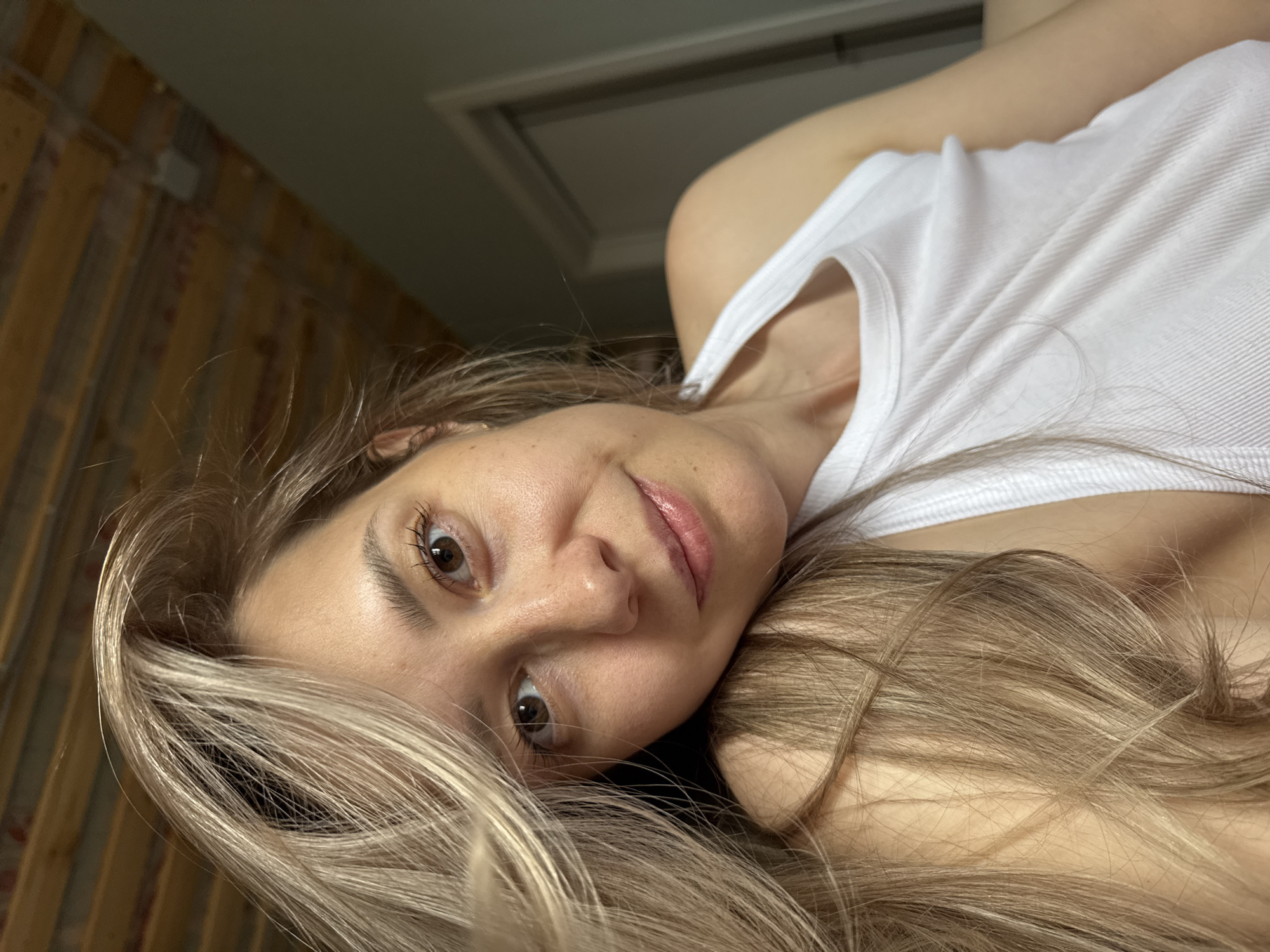 A woman with blonde hair and light skin is lying down, looking directly at the camera. She is wearing a white tank top. The background includes a wooden wall and a ceiling with a rectangular access panel. The location is in a building with wood paneling, but cannot be definitively said to be in the city of Chaikovsky, Russia.
